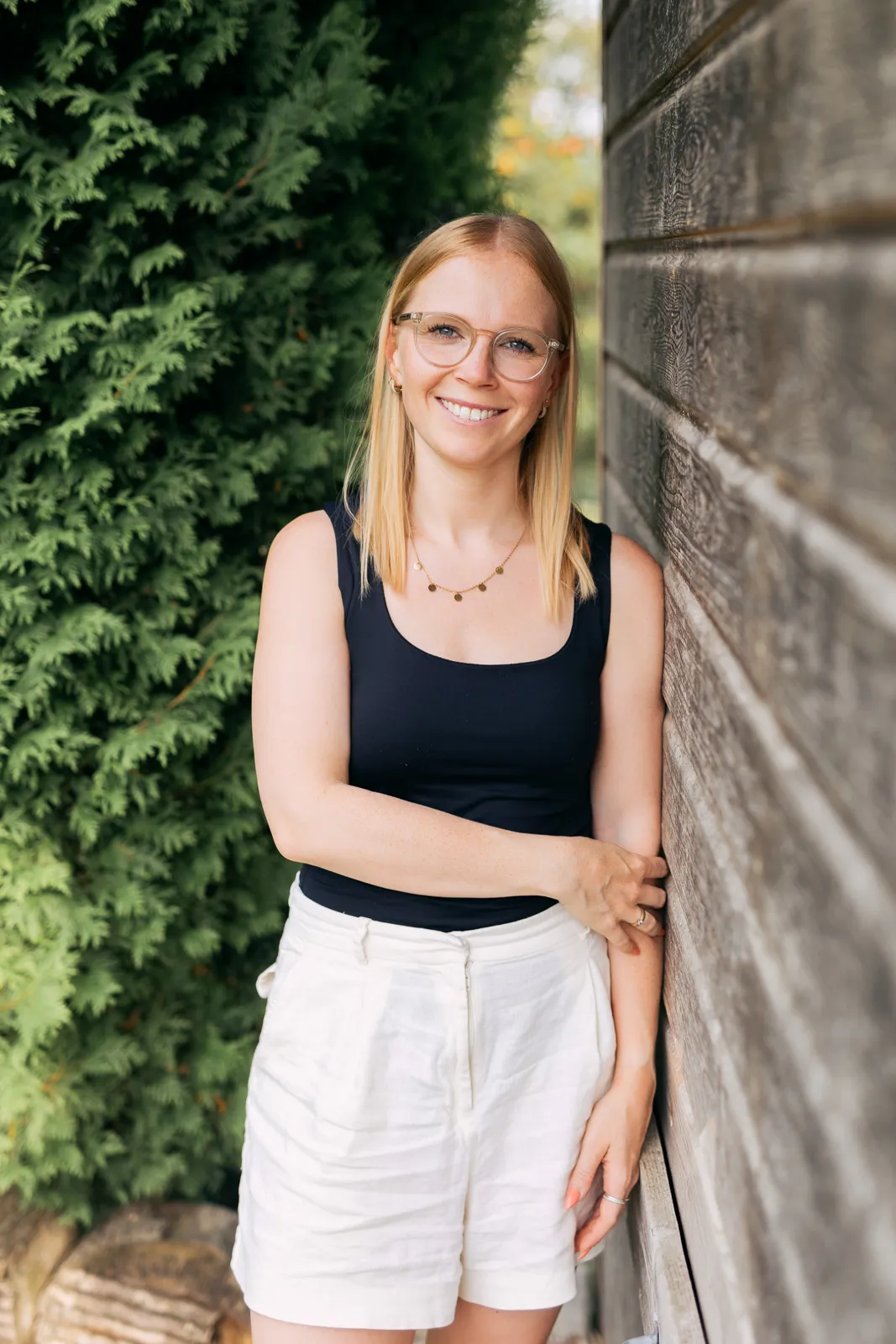 Frau lehnt entspannt am Holzhaus, trägt schwarze Bluse und helle Shorts, natürliches Lächeln für Ringana Produktberatung.