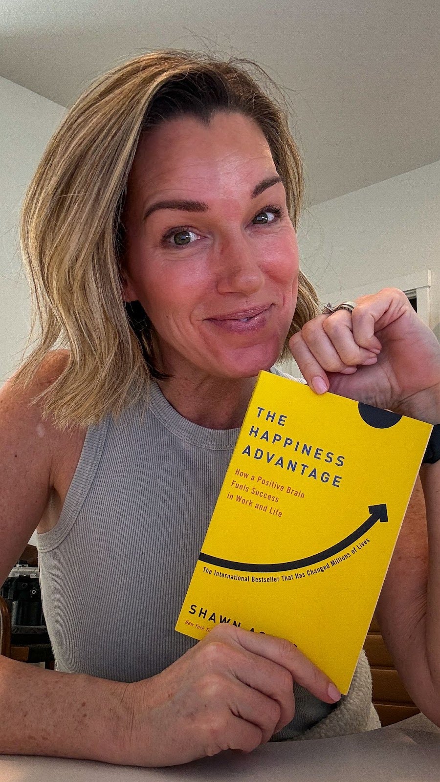 Midlife woman smiling and holding The Happiness Advantage book, promoting positive mindset and success.