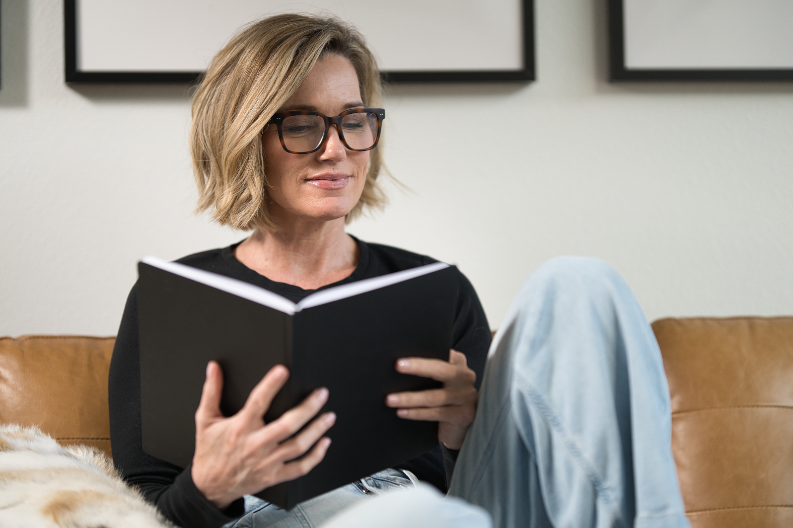 Midlife woman over 40 wearing glasses sitting on a couch, reading a personal development book about growth and wellness