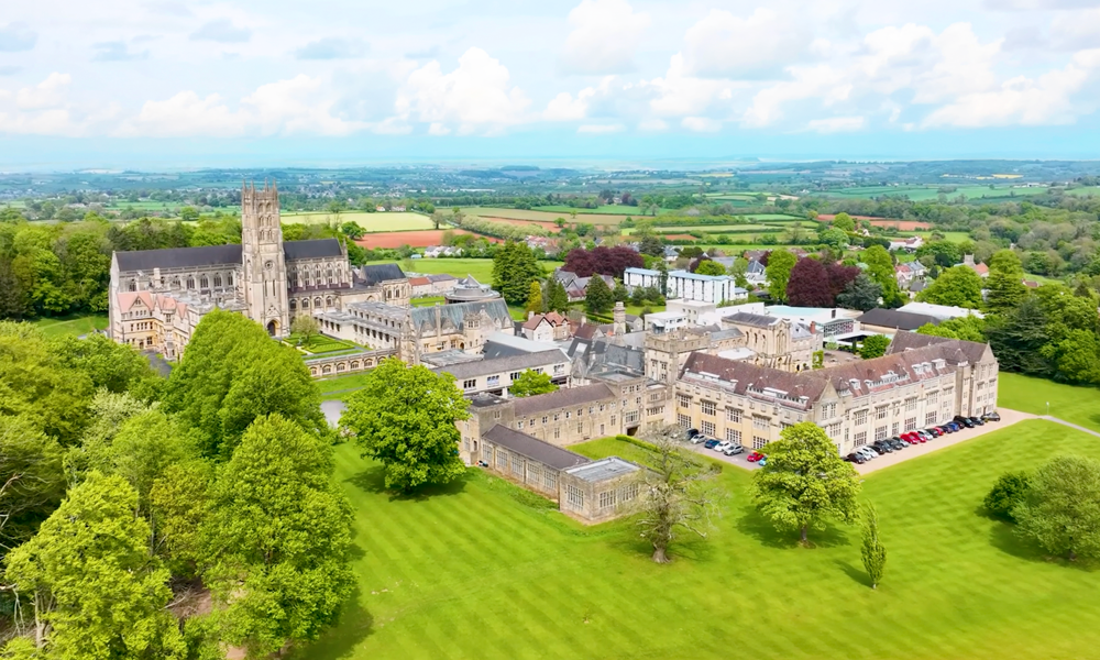 Aerial view of historic stone buildings and lush green lawns surrounded by trees under a blue sky.