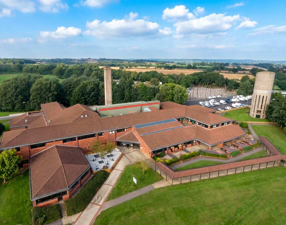 Aerial view of a large red brick building with a courtyard, paths, parking, water tower, and surrounding greenery.