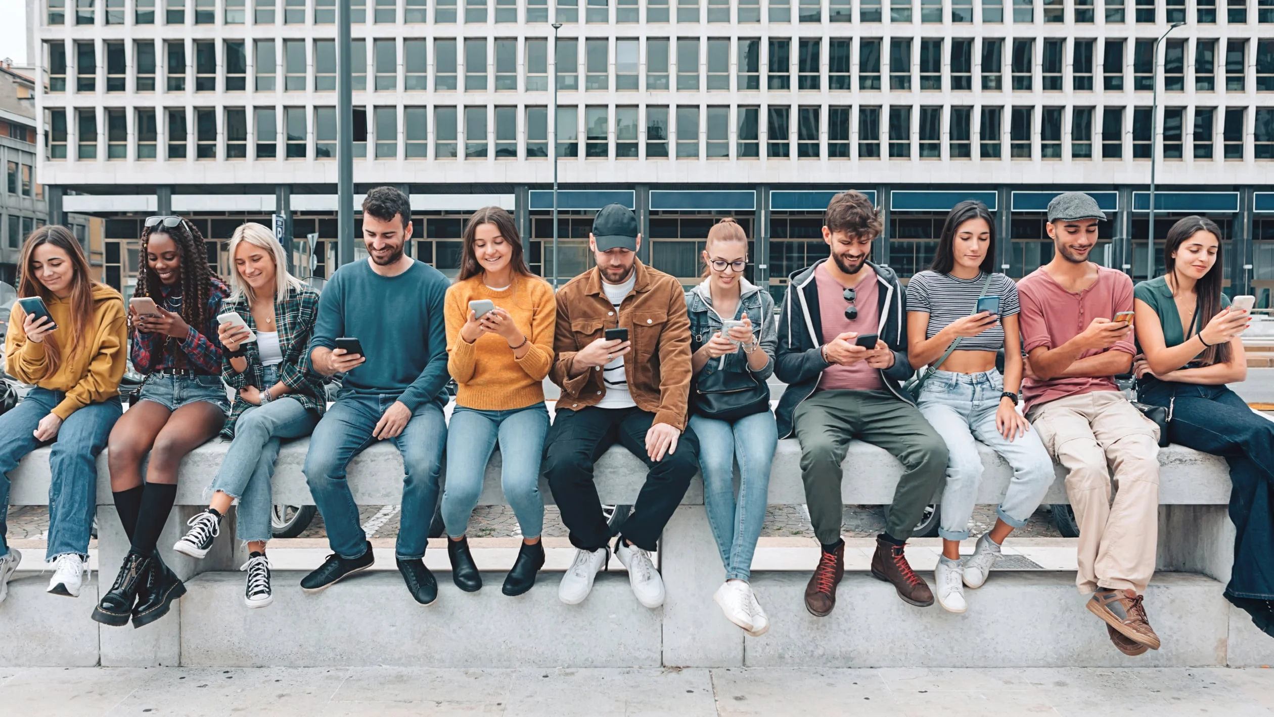A group of eleven young adults sit in a row outdoors, each looking at their smartphones, with a modern office building in the background.