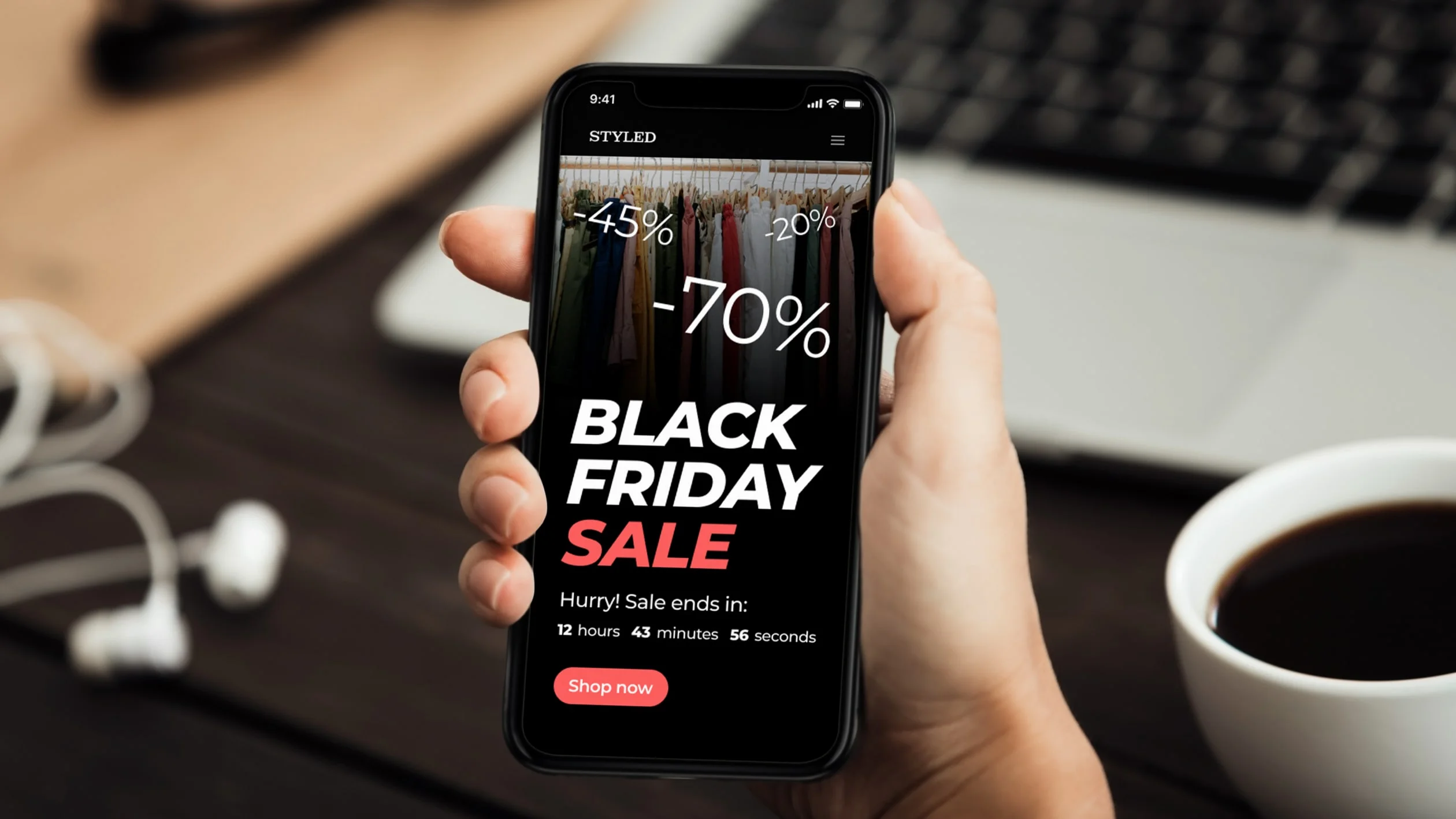 A hand holds a smartphone displaying a clothing website with a Black Friday Sale ad showing discounts of -45%, -20%, and -70%, along with a countdown timer and Shop now button. A laptop and coffee cup are in the background.