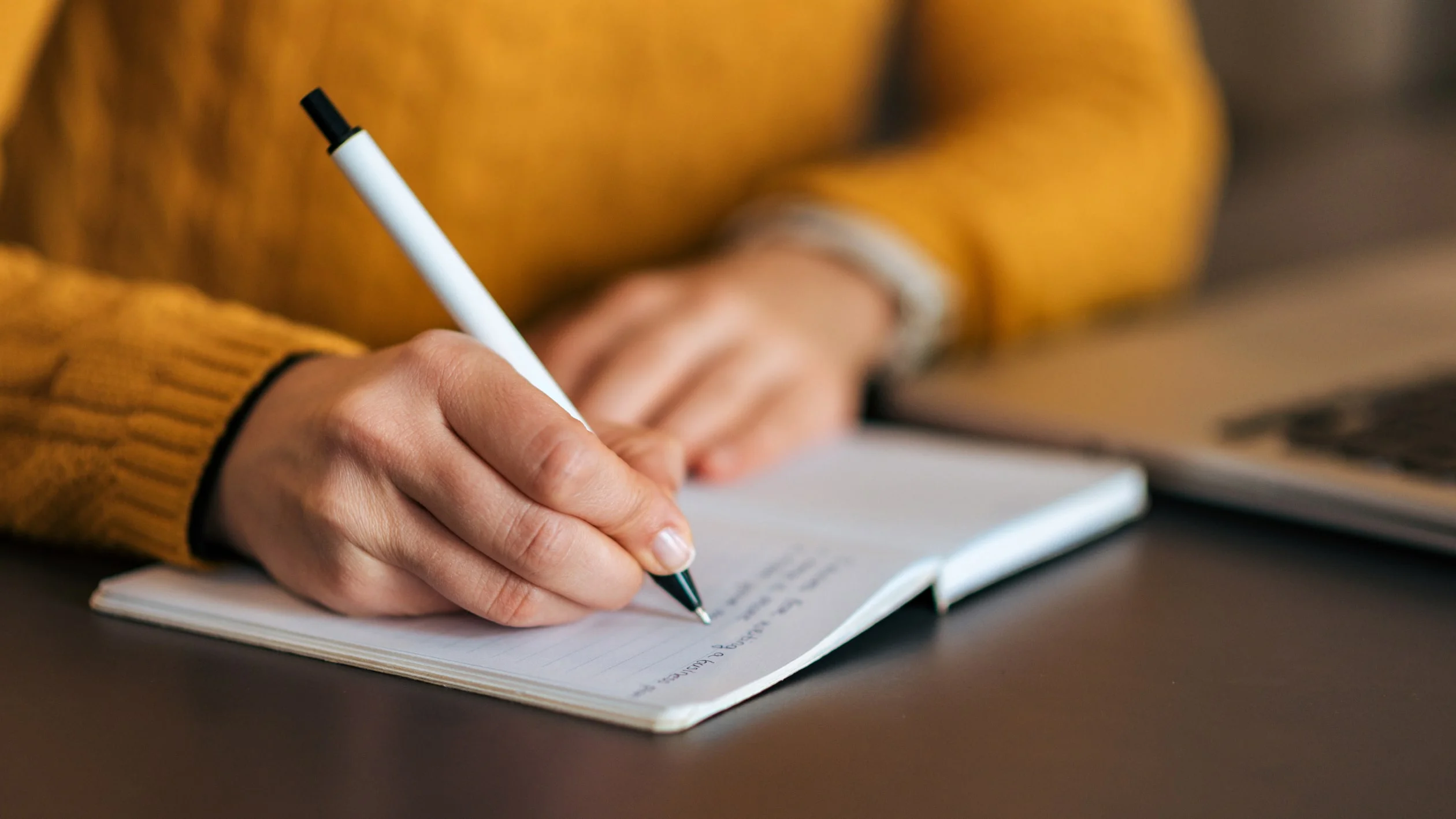 Close-up of a person wearing a yellow sweater writing in a notebook with a black and white pen, with a laptop partially visible on the desk in the background.