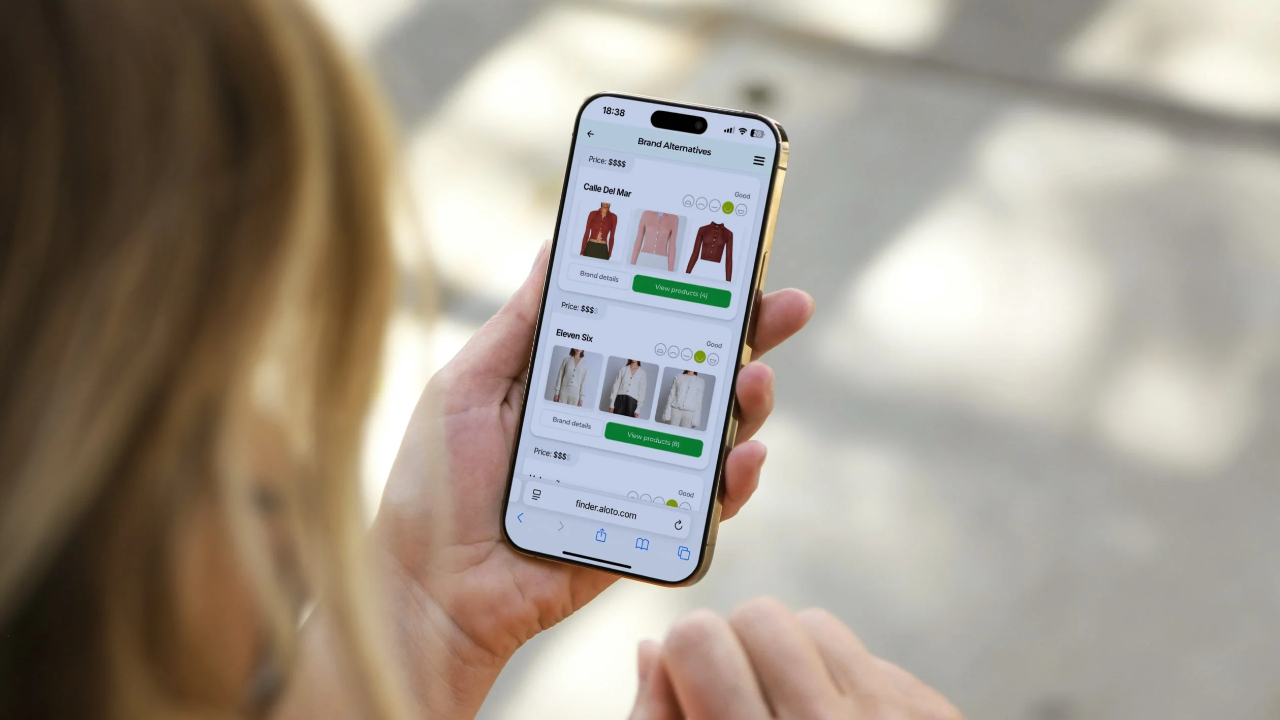 A person holds a smartphone displaying an online clothing store with images and prices of various tops and dresses, while standing outdoors on a sunny day.