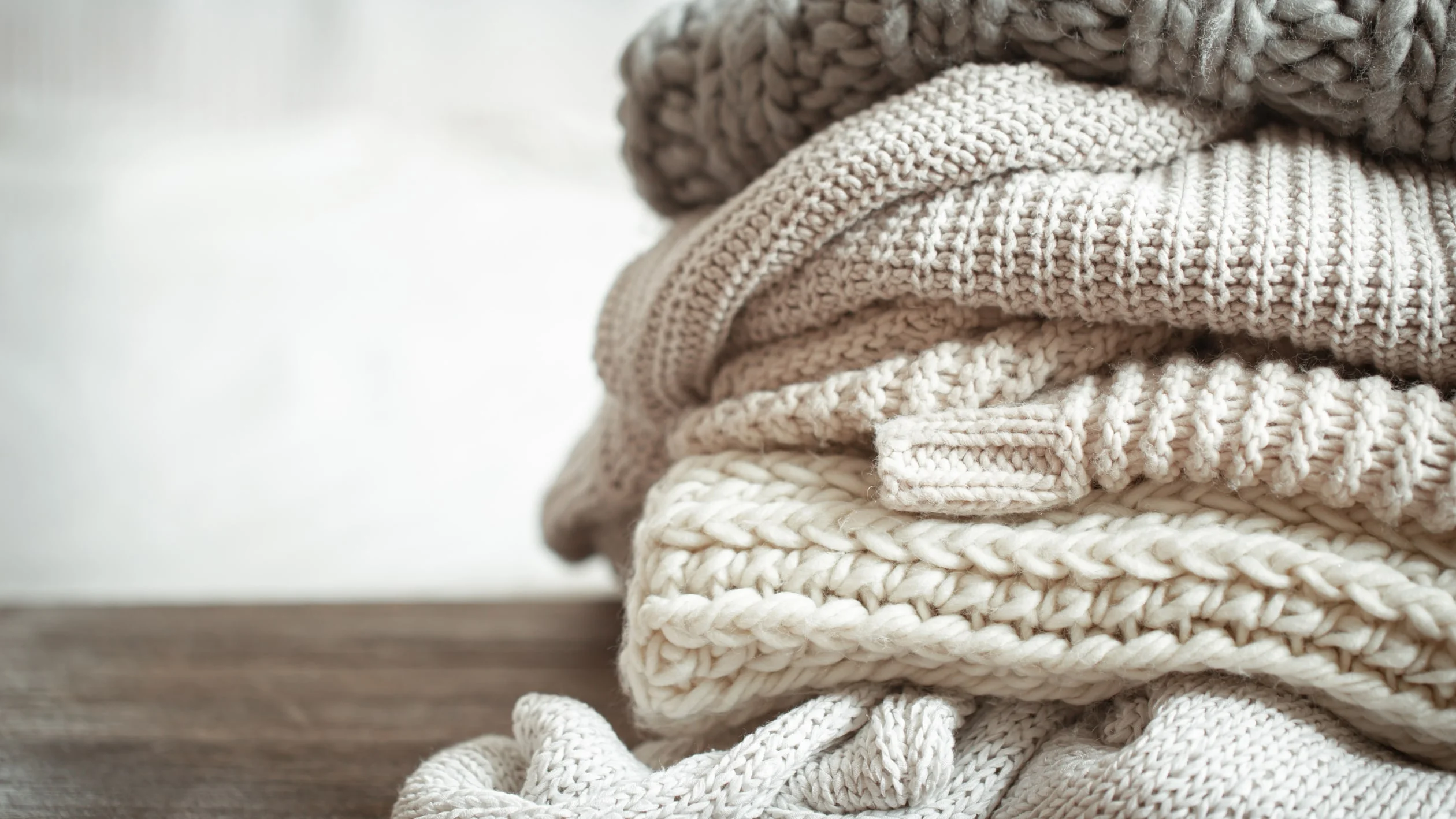 A stack of cozy, knitted sweaters in neutral tones, neatly folded on a wooden surface with a soft, blurred background.