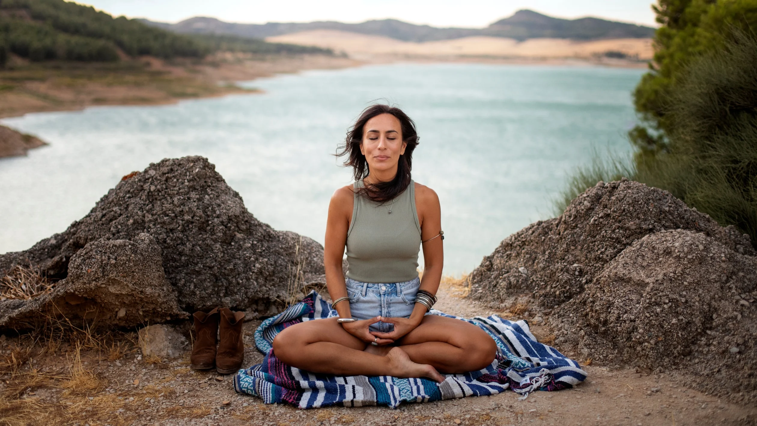 A woman sits cross-legged on a blanket, meditating outdoors by a lake with mountains in the background. She is wearing a tank top and shorts, and her eyes are closed, surrounded by rocks and nature.