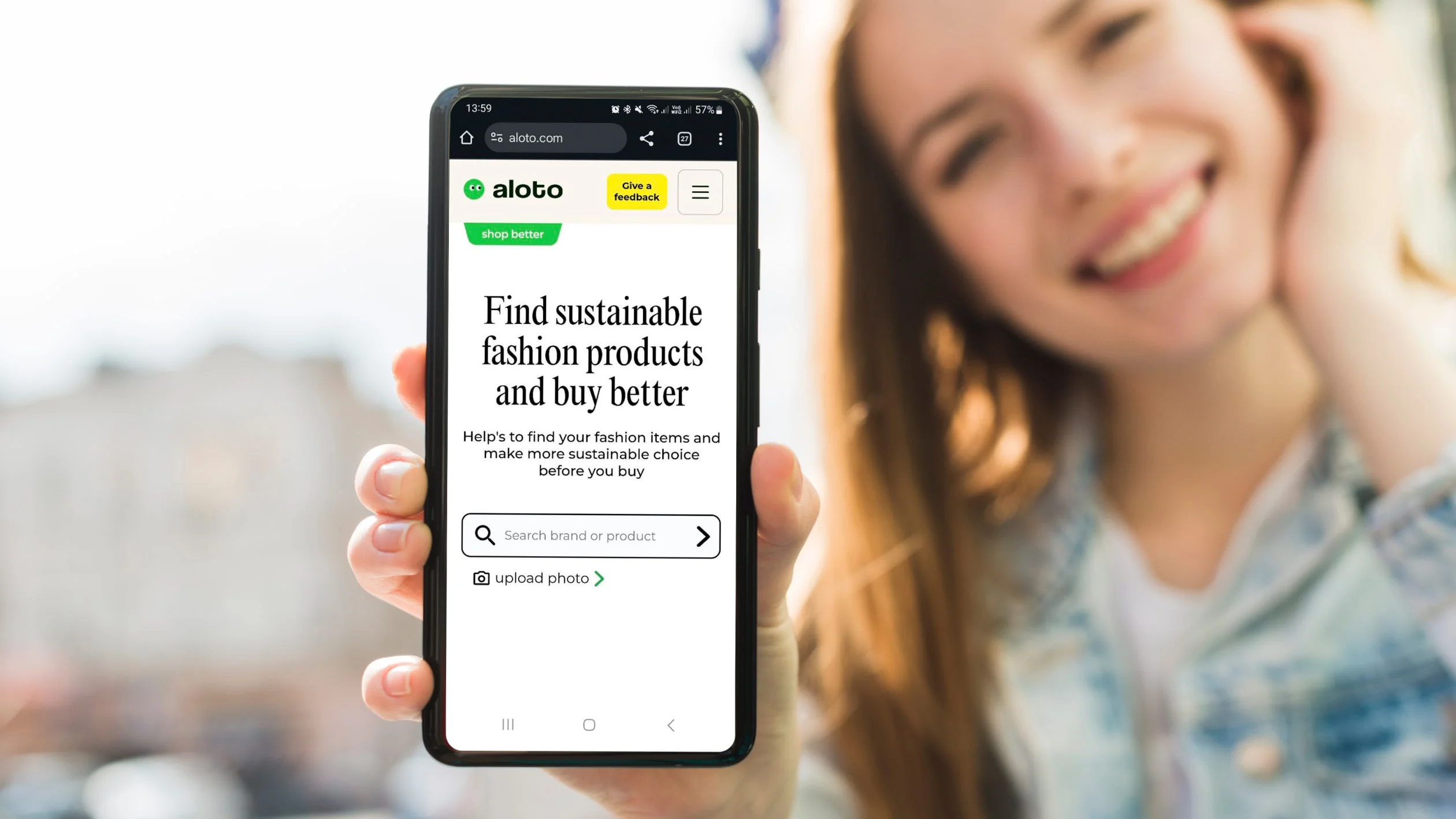 A smiling person holds a smartphone displaying a website called aloto with the text: Find sustainable fashion products and buy better and a search bar for products. The background is blurred.