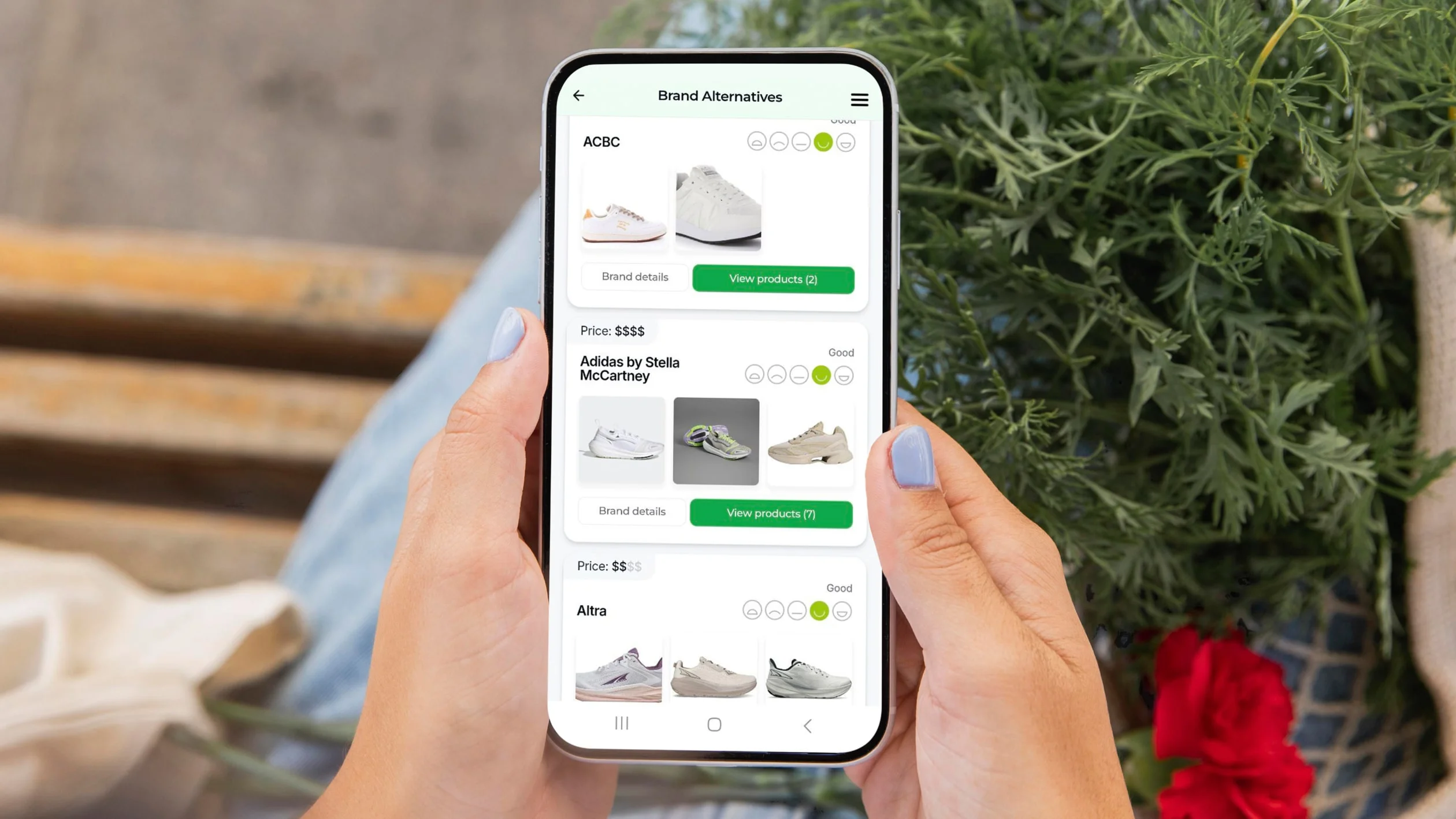A person holds a smartphone displaying a shopping app with sneaker alternatives, prices, and ratings. Plants and a red flower are visible in the background.