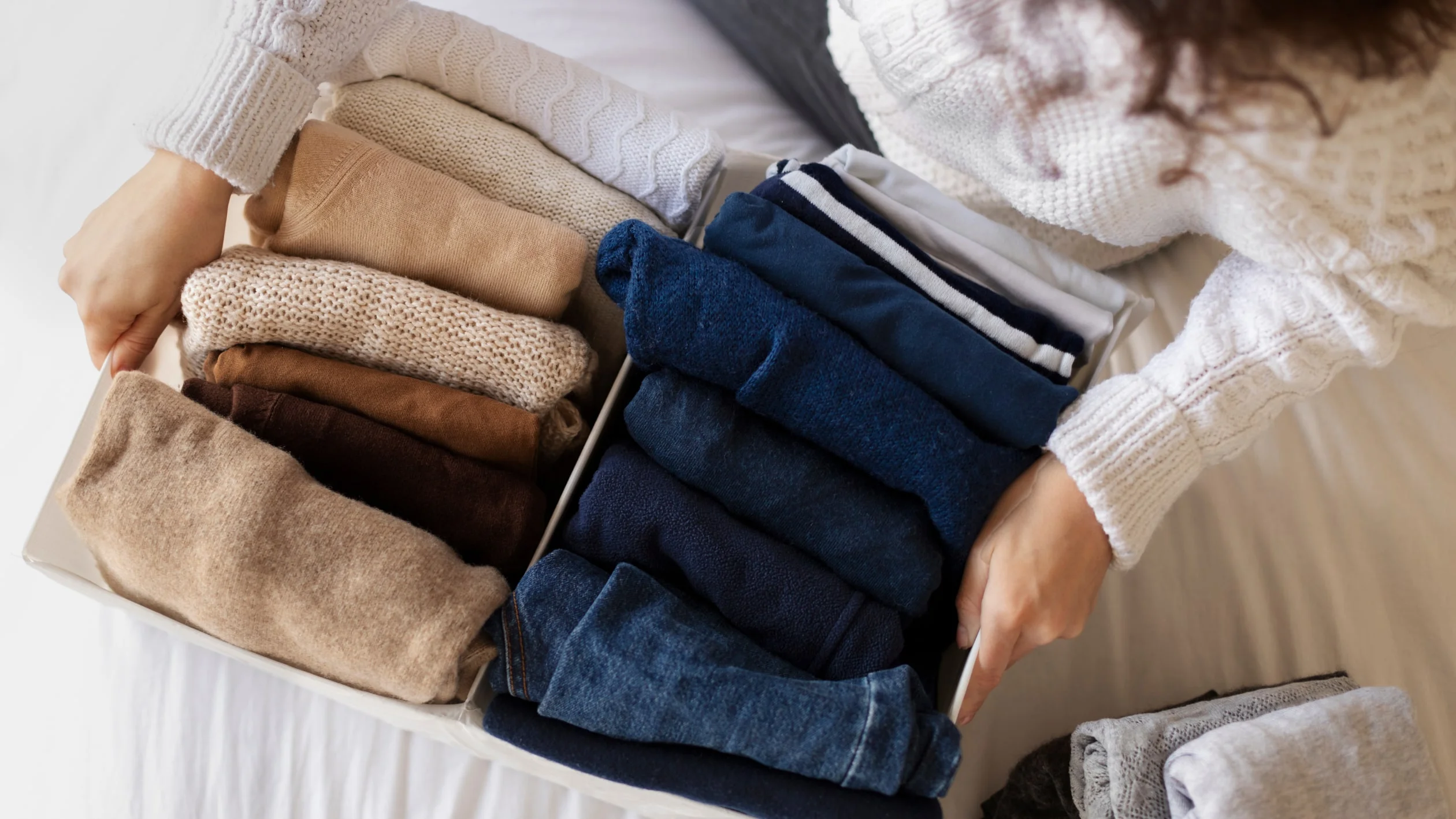 A person in a white sweater organizes a drawer of neatly folded clothes in shades of blue, brown, and beige, conveying a sense of order and calm.