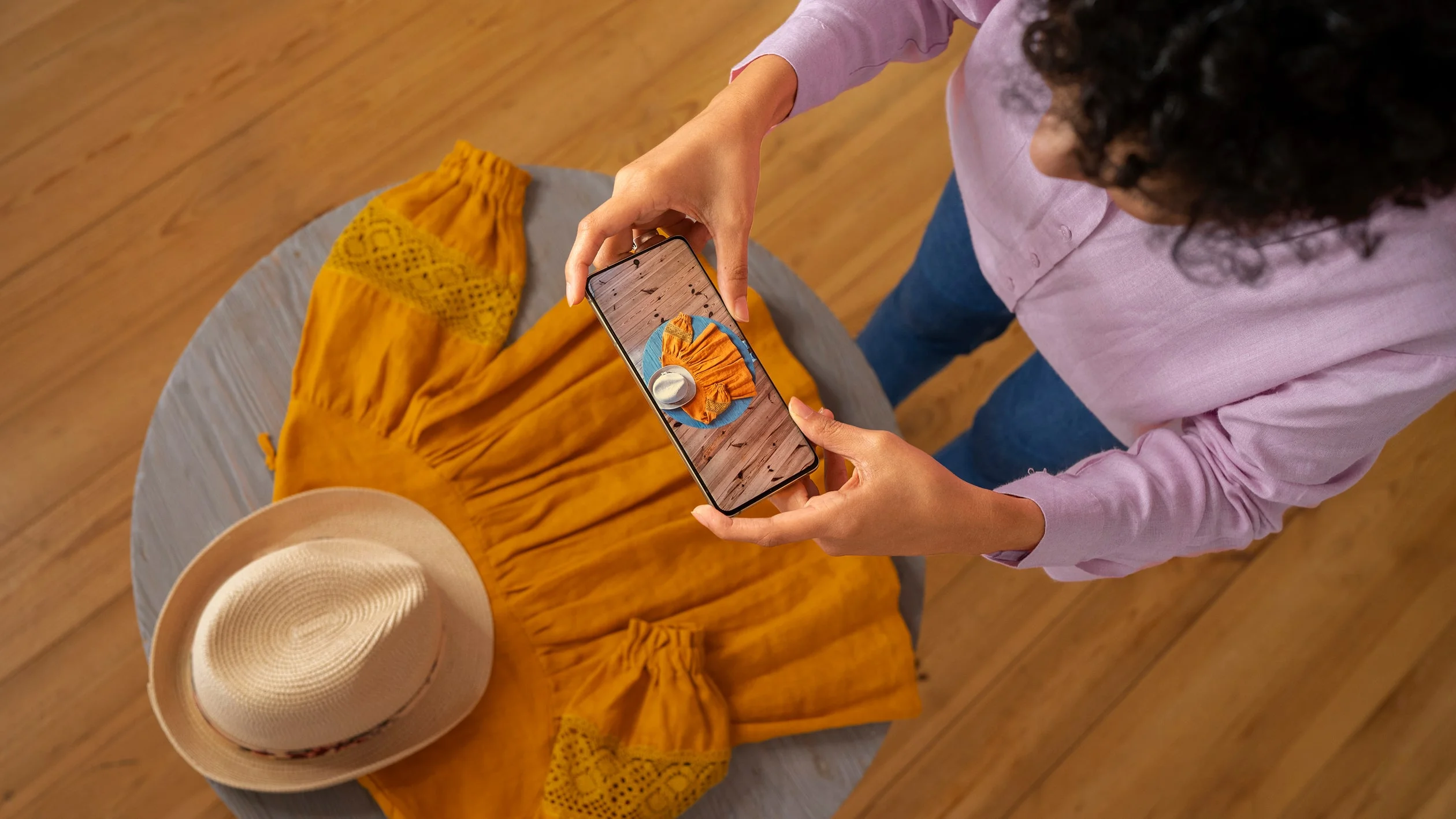 A person in a pink shirt and jeans photographs an orange dress with lace sleeves on a round table. A hat lies beside it, conveying a casual, creative vibe.