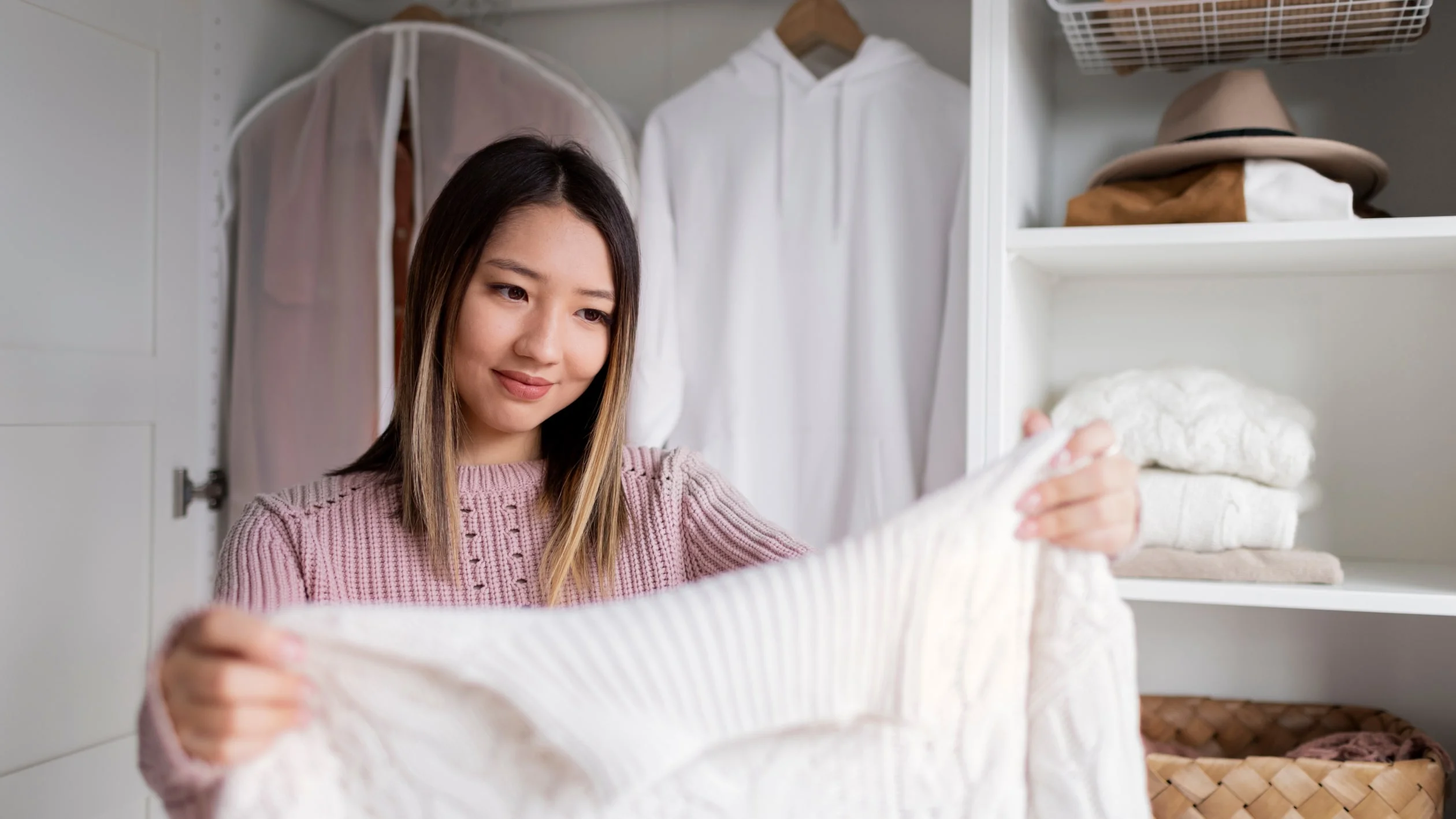 A woman in a pink sweater examines a white garment with a thoughtful expression in a tidy closet. The closet has neatly folded clothes and hats.