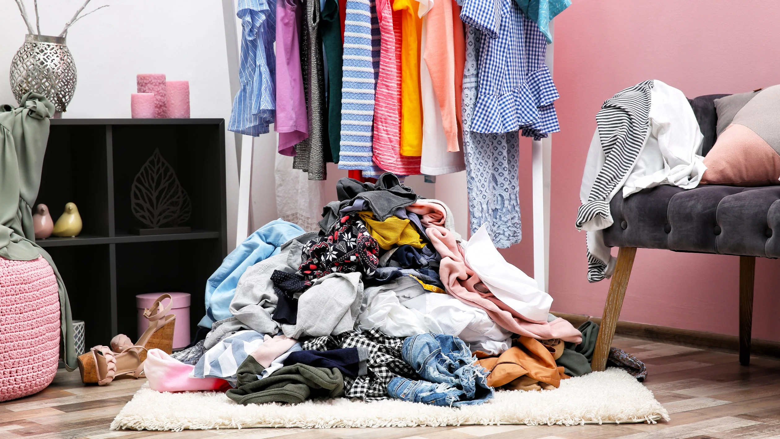 A cluttered room with a large pile of mixed clothing on a white rug. Colorful clothes hang on a rack. The scene looks disorganized yet lively.