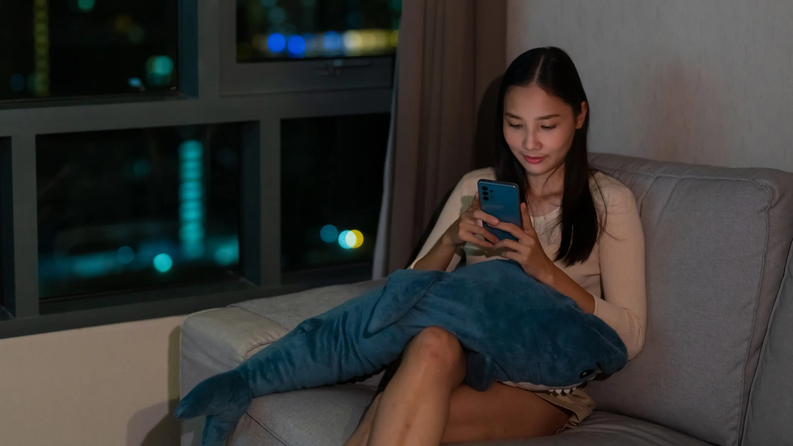 A woman sitting on a couch, holding a phone in her hand, appears engaged in conversation or browsing content.