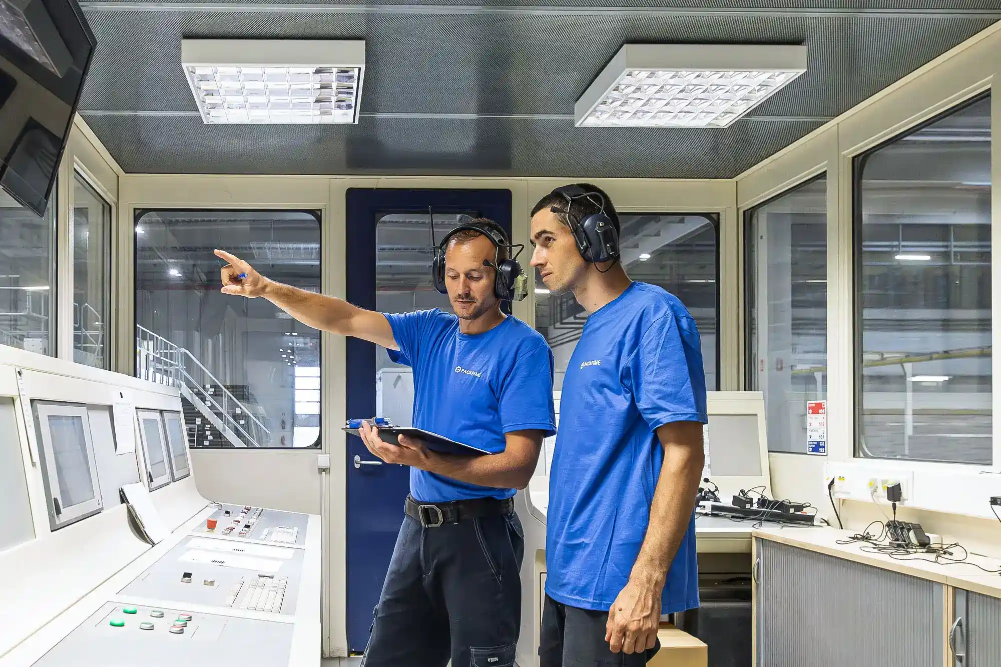two workers in headsets, pointing at the machine, wearing branded shirts