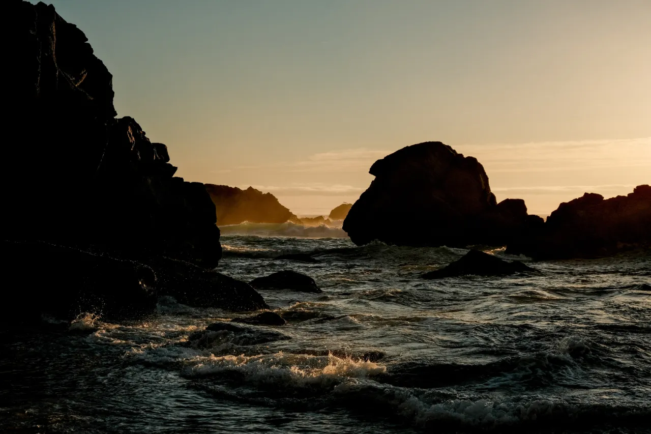 Ocean waves crashing against dark rocky cliffs at sunset with an orange sky.