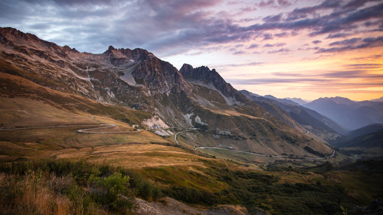 Winding mountain road cutting through rugged peaks and grassy hills under a colorful sunset sky.