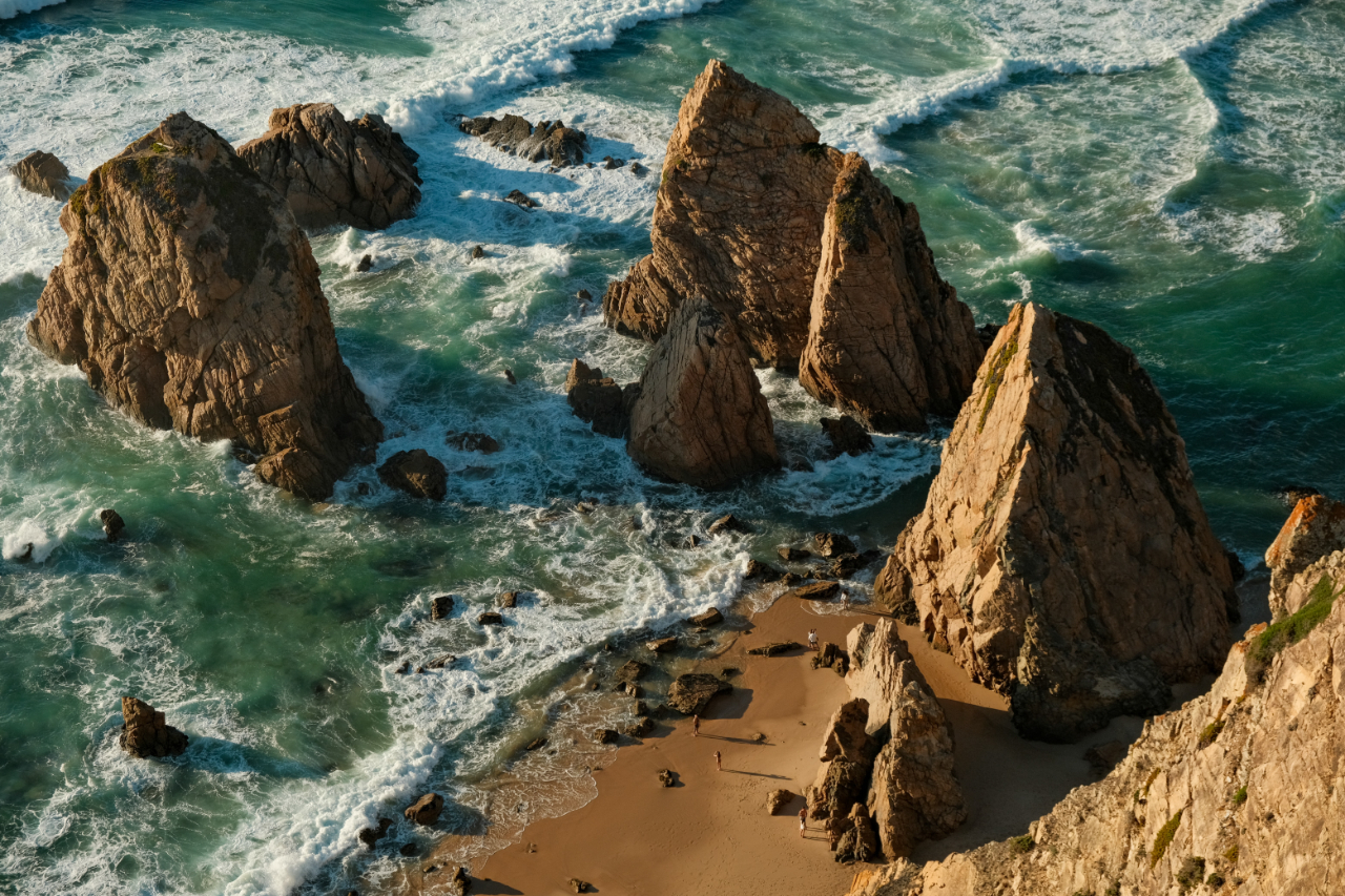 Rock formations rising from turquoise ocean waves next to a sandy beach.