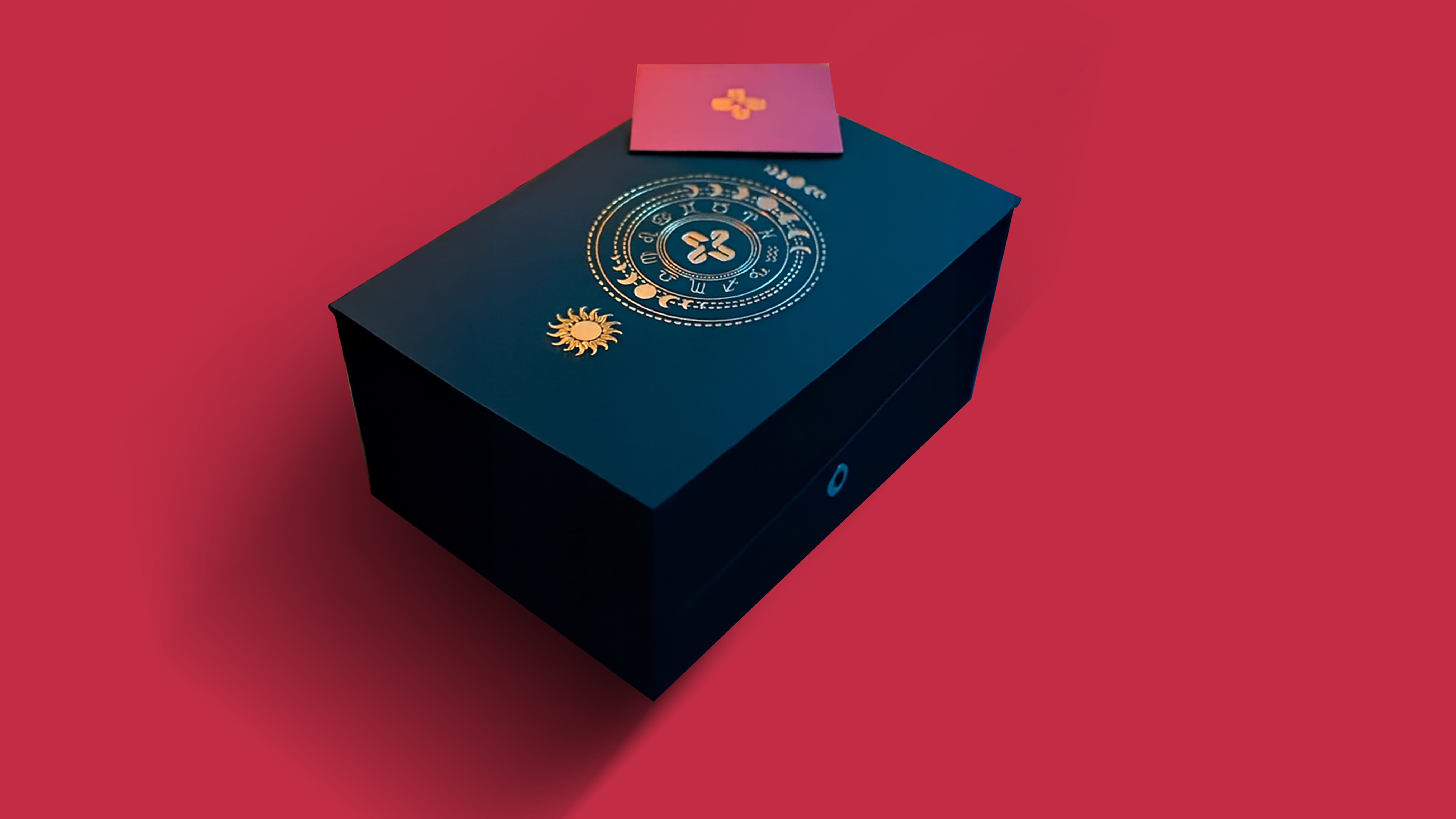 Black rectangular box with a gold sun and zodiac wheel design on top, set against a red background with a pink card bearing a gold symbol placed on it.