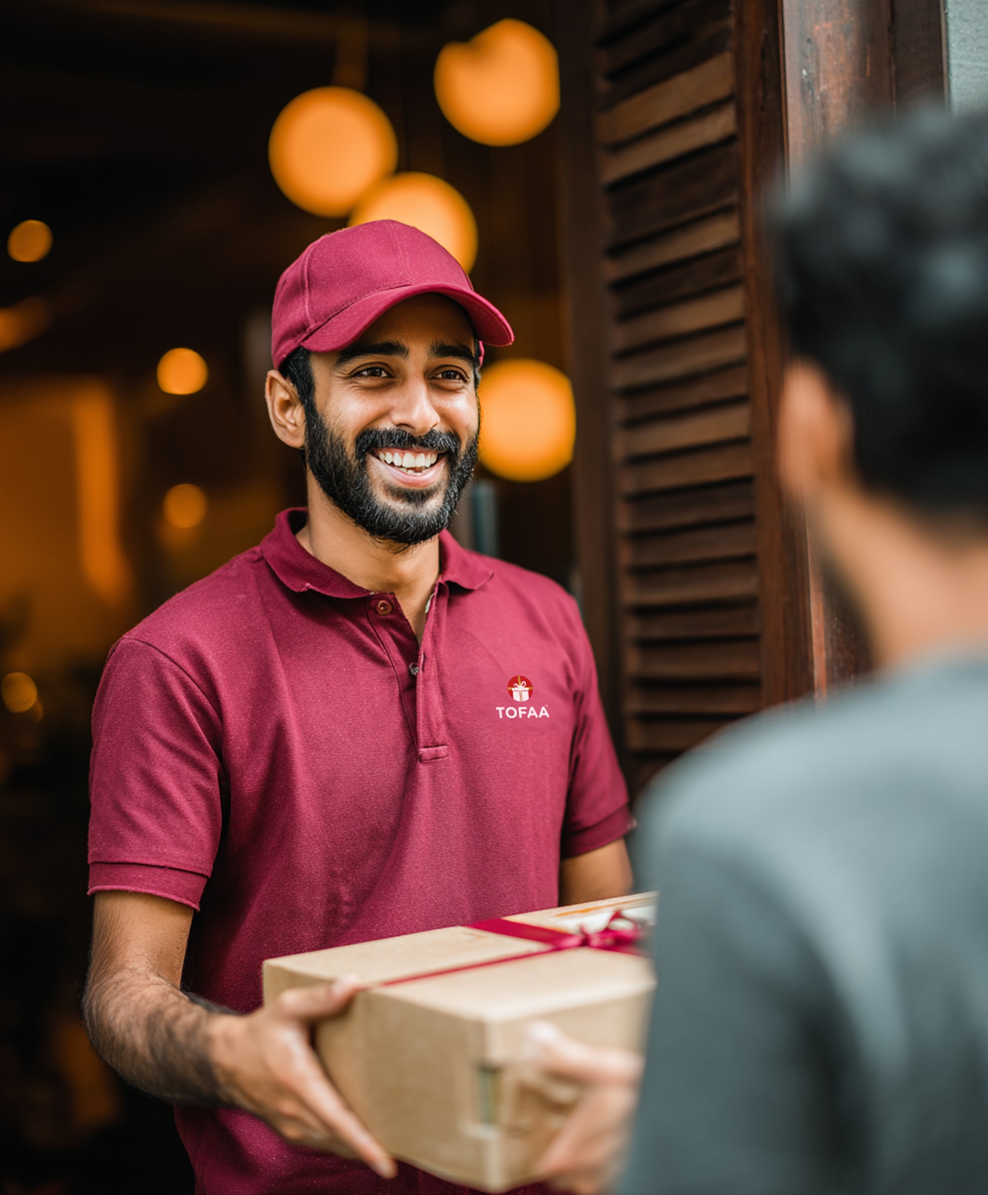 Smiling delivery man in a maroon uniform handing a wrapped gift box to a customer at a door.