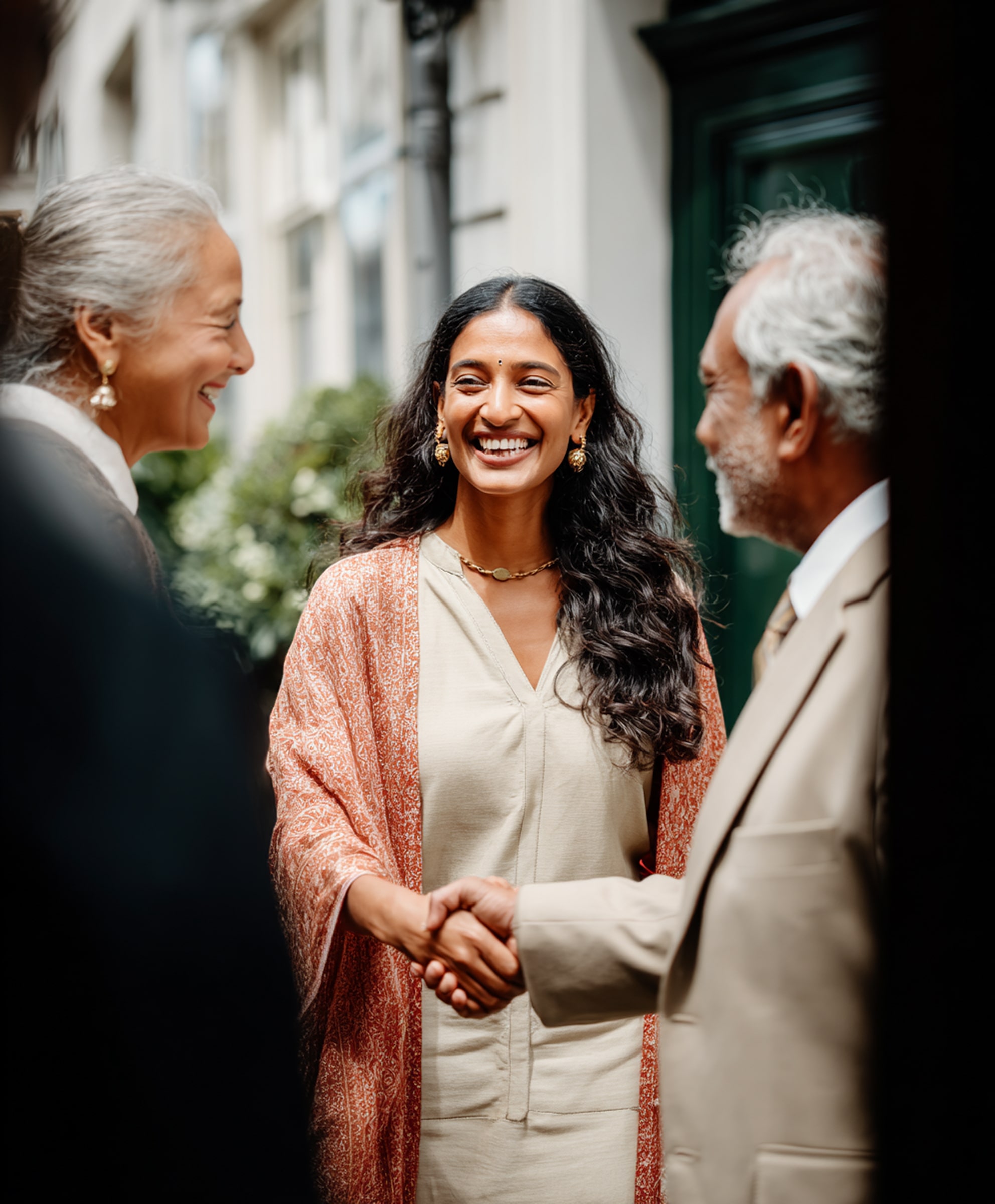 Smiling young woman wearing an orange shawl shaking hands with an older man in a beige suit while an older woman looks on happily.