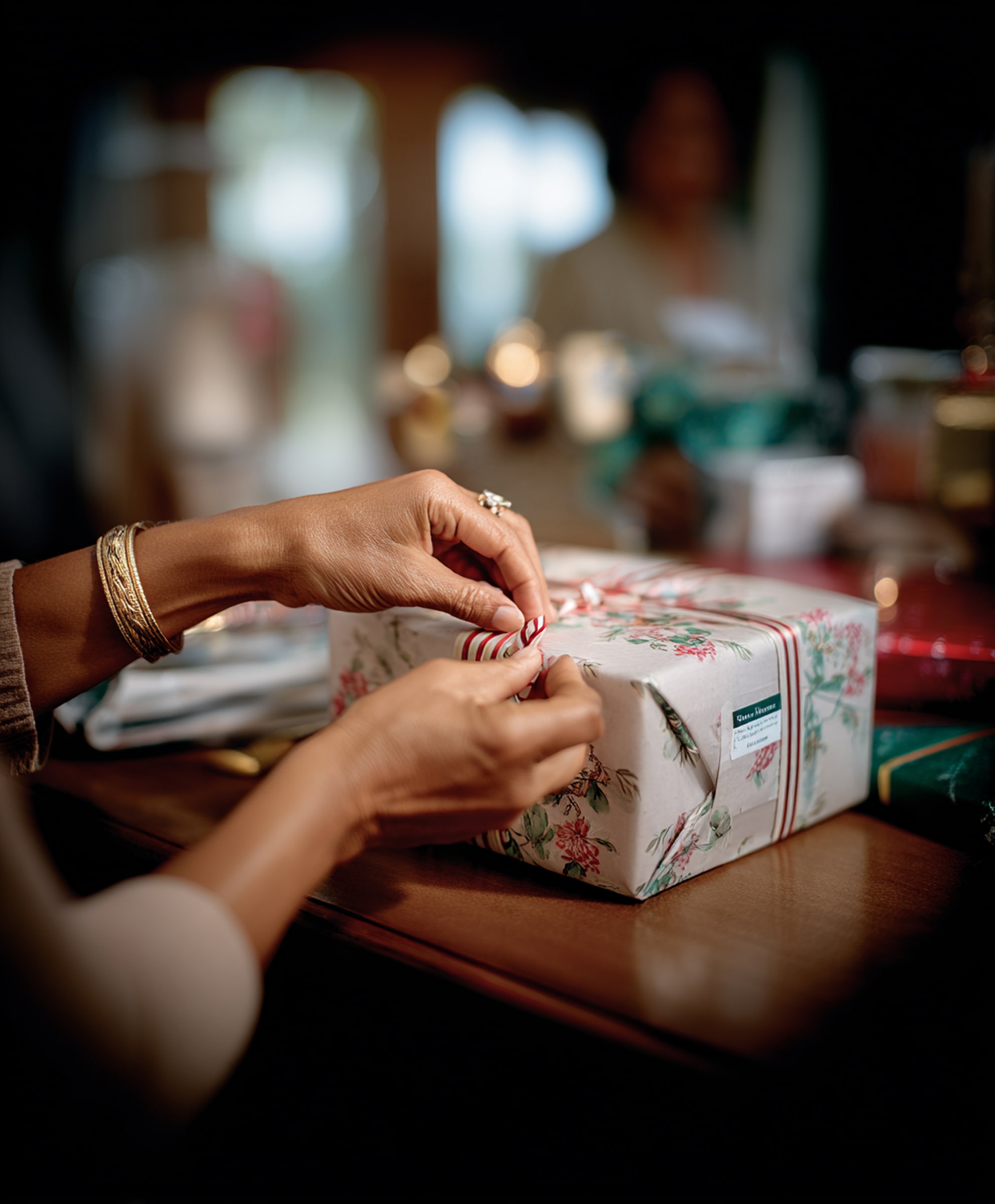 Hands tying a red and white striped ribbon on a floral wrapped gift box on a wooden table.