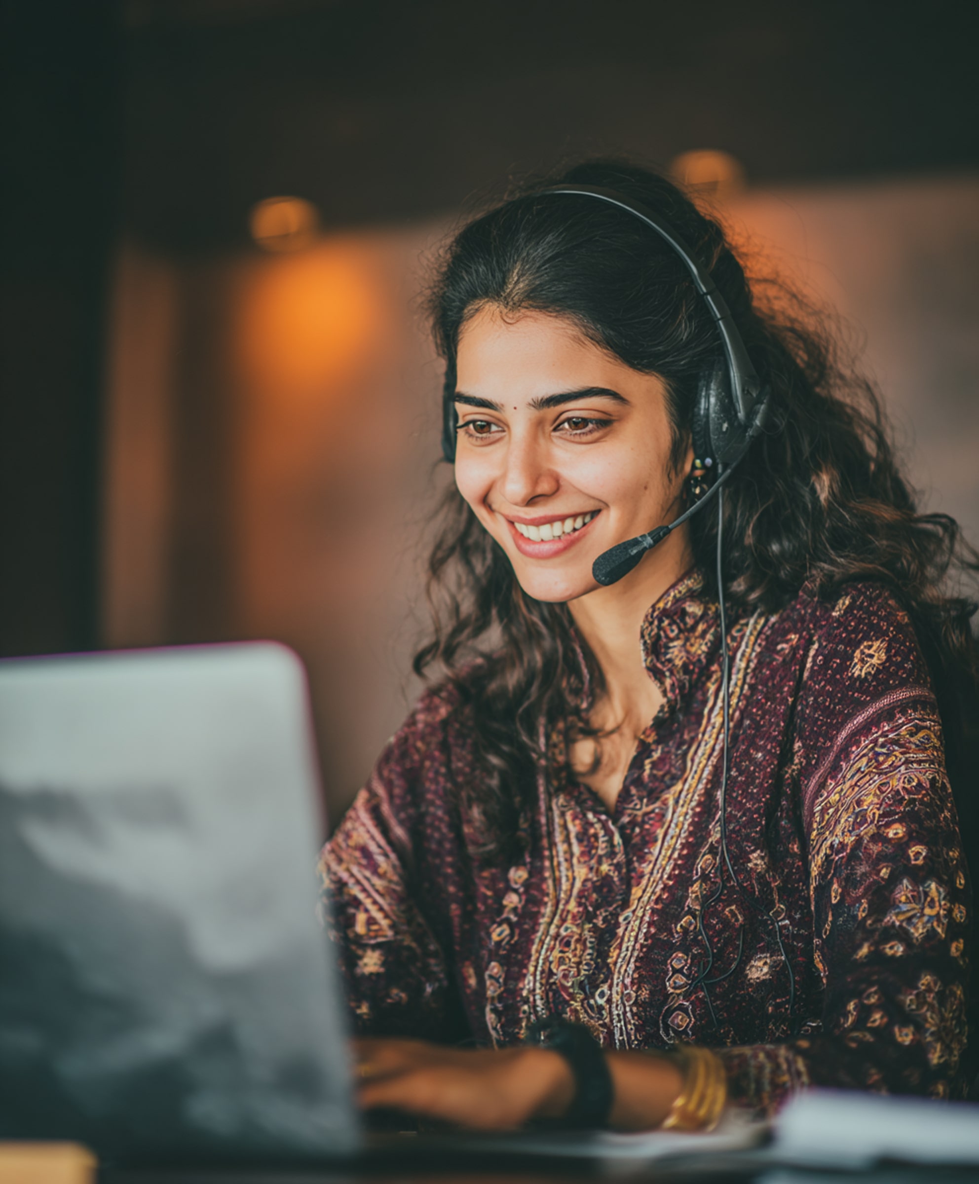 Smiling woman with headset working on a laptop in a softly lit room.