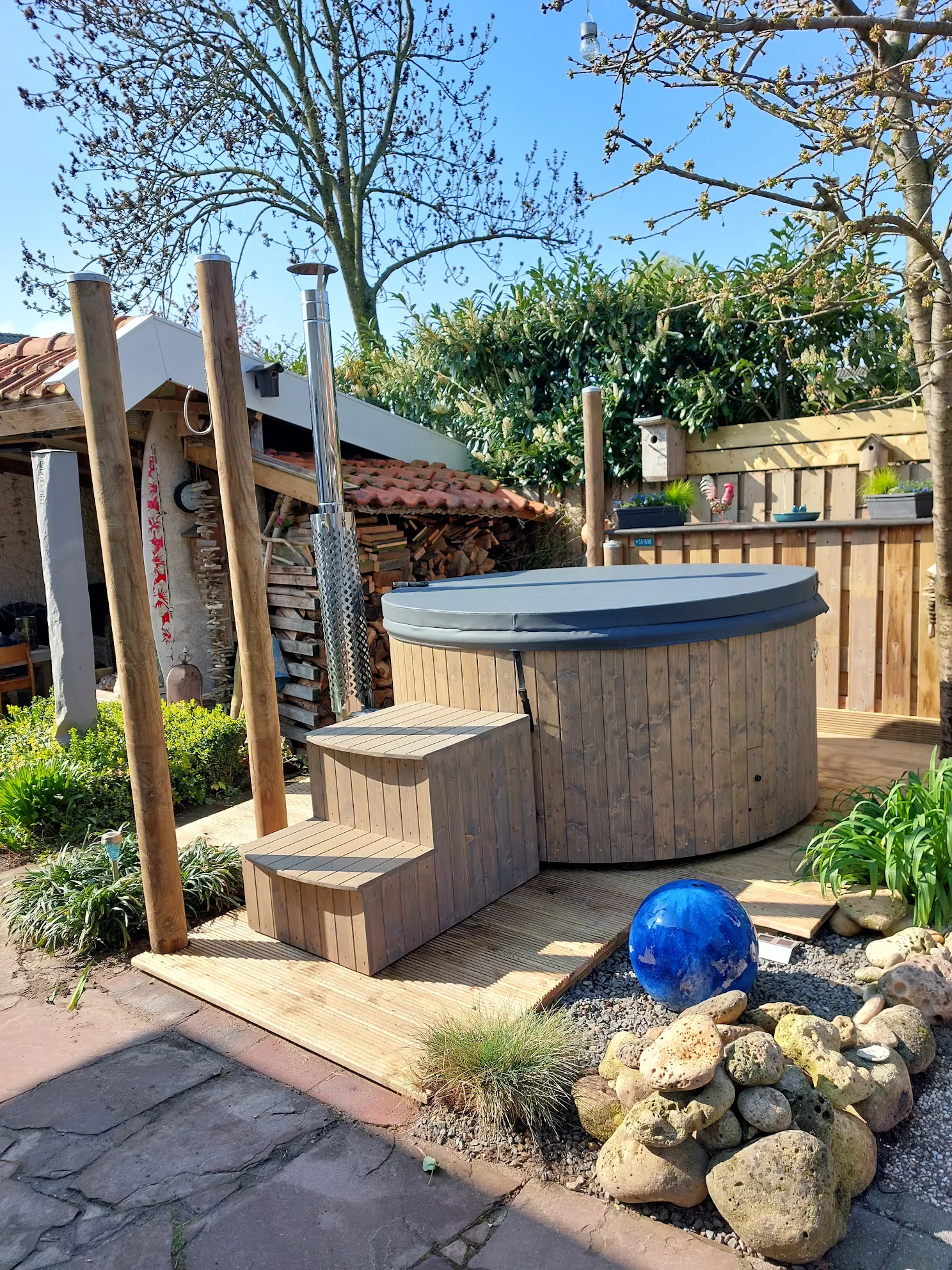 Ronde houten hottub met grijze cover, houten opstap en houtgestookte kachel in een zonnige tuin met sierstenen en blauwe glazen bol