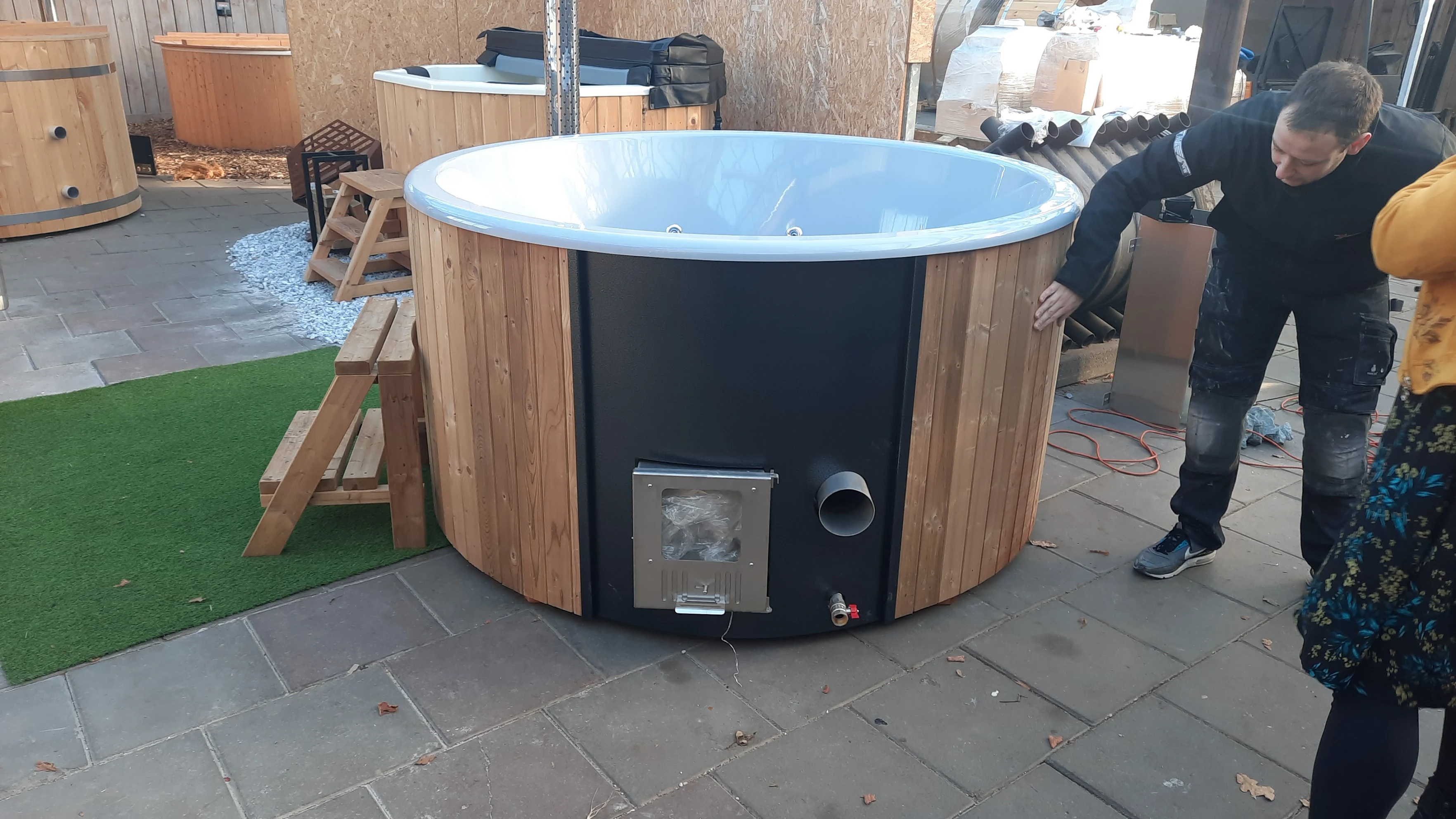 Houten hottub met witte kunststof binnenkant, voorzien van een zwarte geïntegreerde buitenkachel en opstap, terwijl een man de afwerking controleert