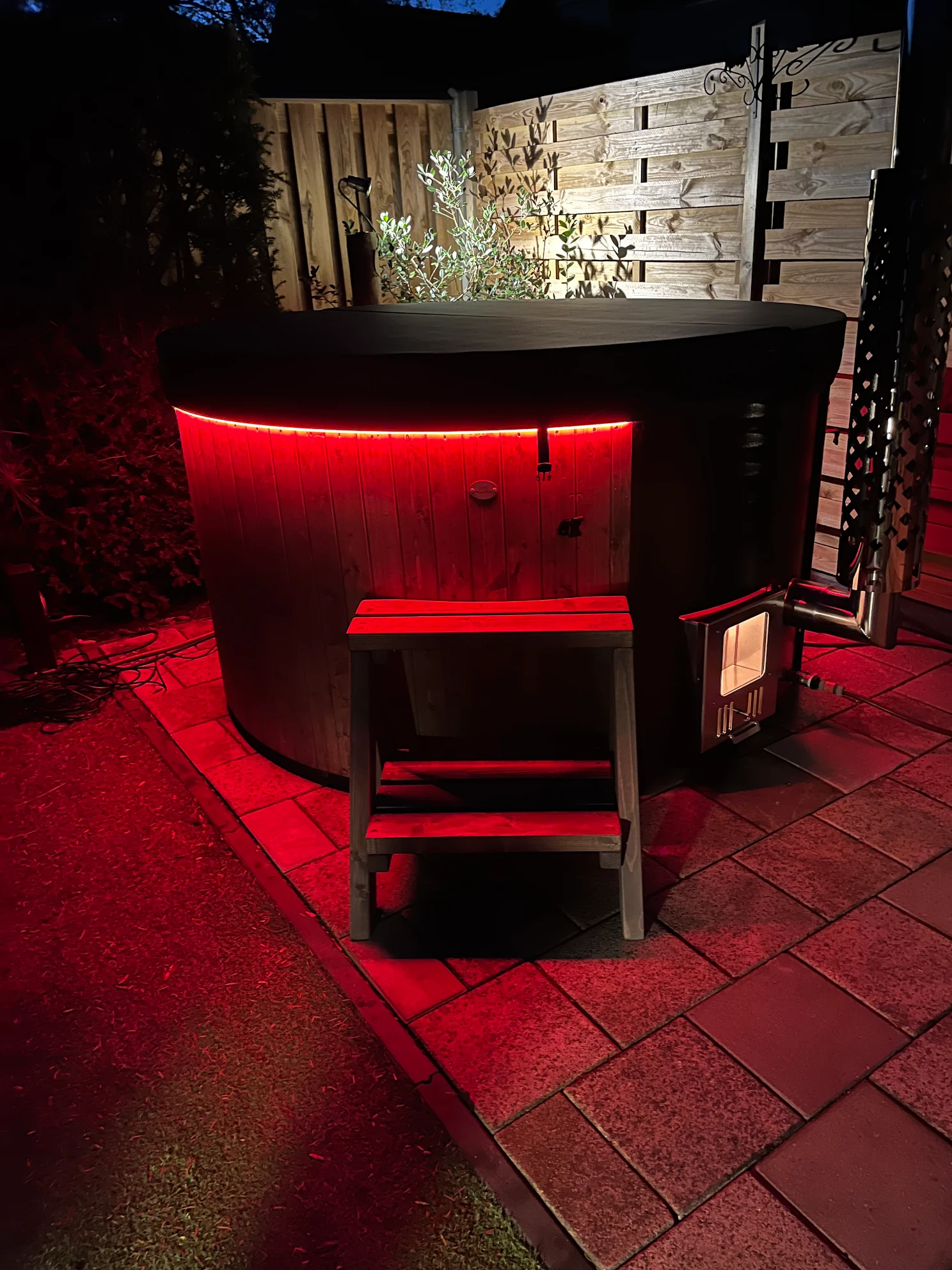 Ronde houten hottub met zwarte afdekking, rode ledverlichting en houtkachel op een betegeld terras in de avond
