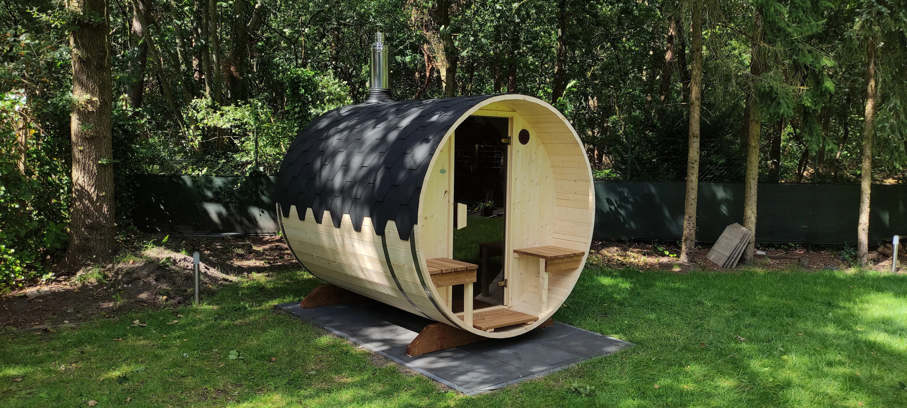 Houten barrelsauna met zwart dak en houten zitjes geplaatst in een bosrijke tuinomgeving