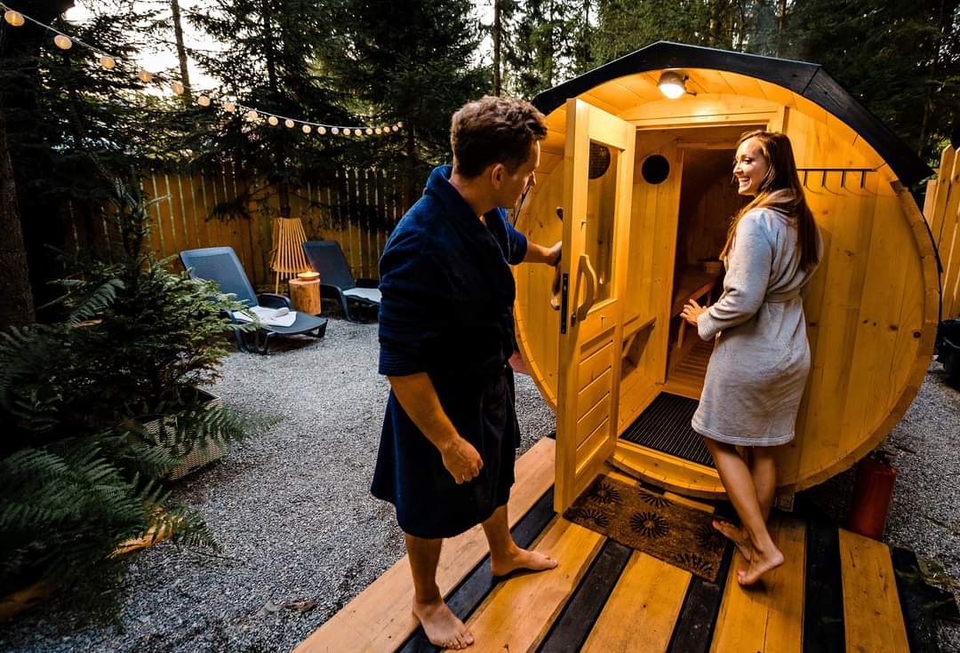 Man en vrouw in badjas stappen een houten tonsauna binnen in een sfeervol verlichte tuin met ligstoelen en bosrijke achtergrond