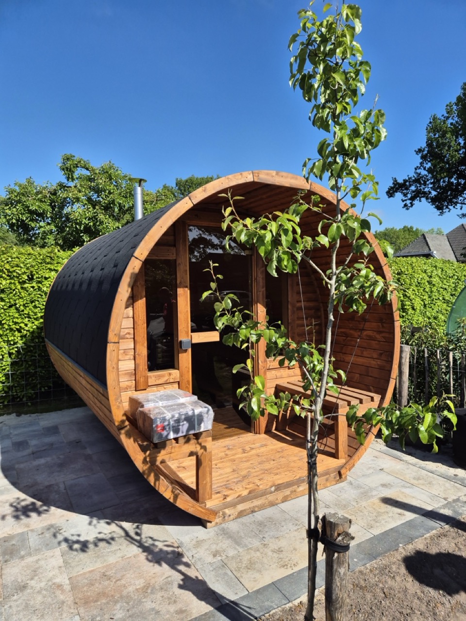Houten barrelsauna met zwart dak, geplaatst op een terras in een groene tuin