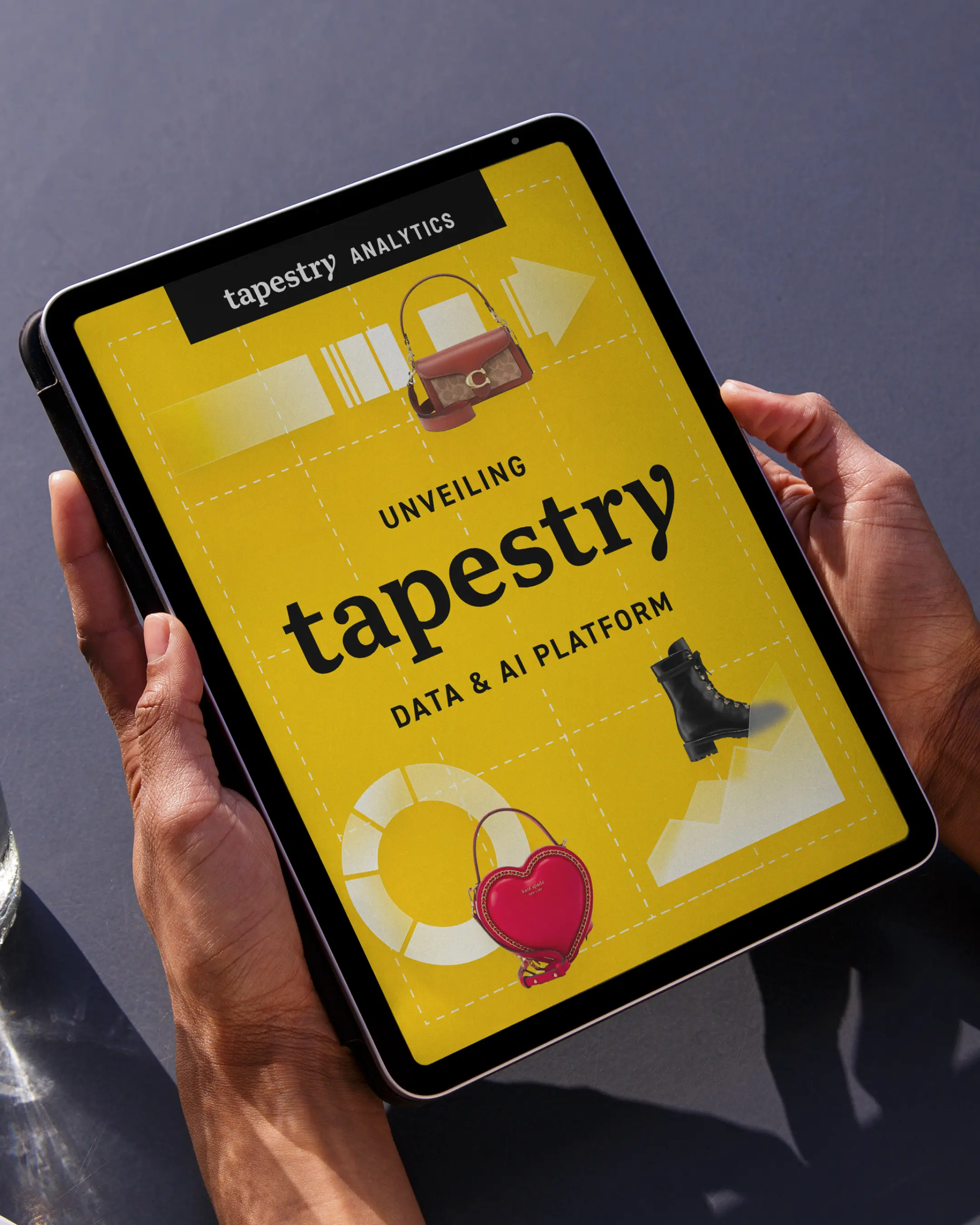 Person holding a tablet showing a yellow screen with 'tapestry Analytics' and 'Unveiling tapestry Data & AI Platform' text, alongside images of a handbag, heart-shaped purse, and black boot.