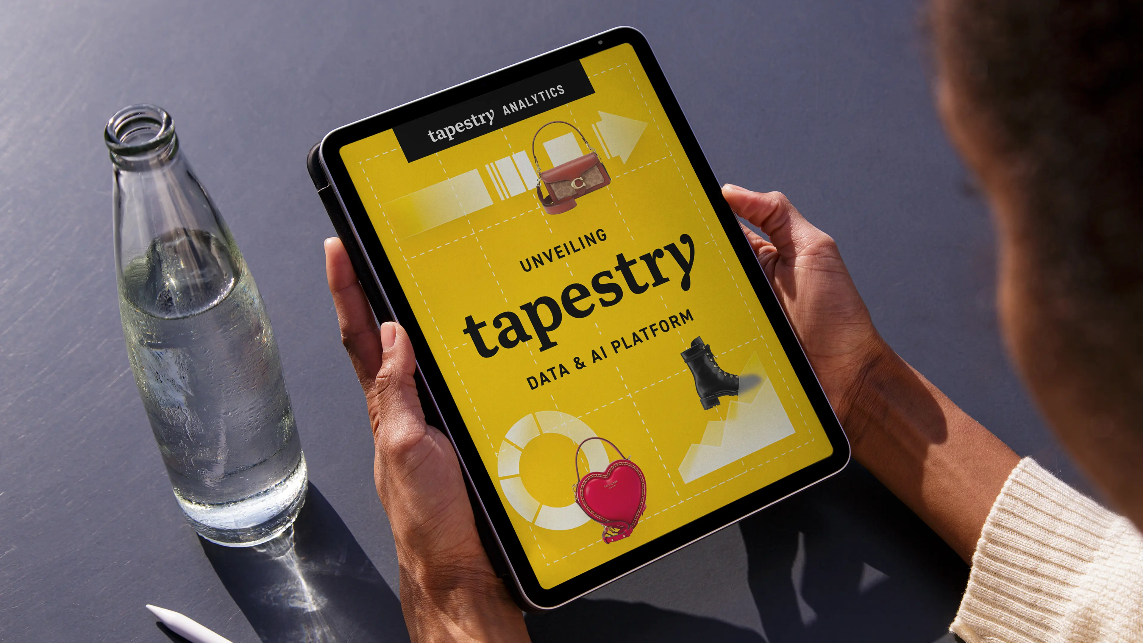 Person holding a tablet showing 'Unveiling tapestry Data & AI Platform' with images of handbags and a boot on a yellow background.