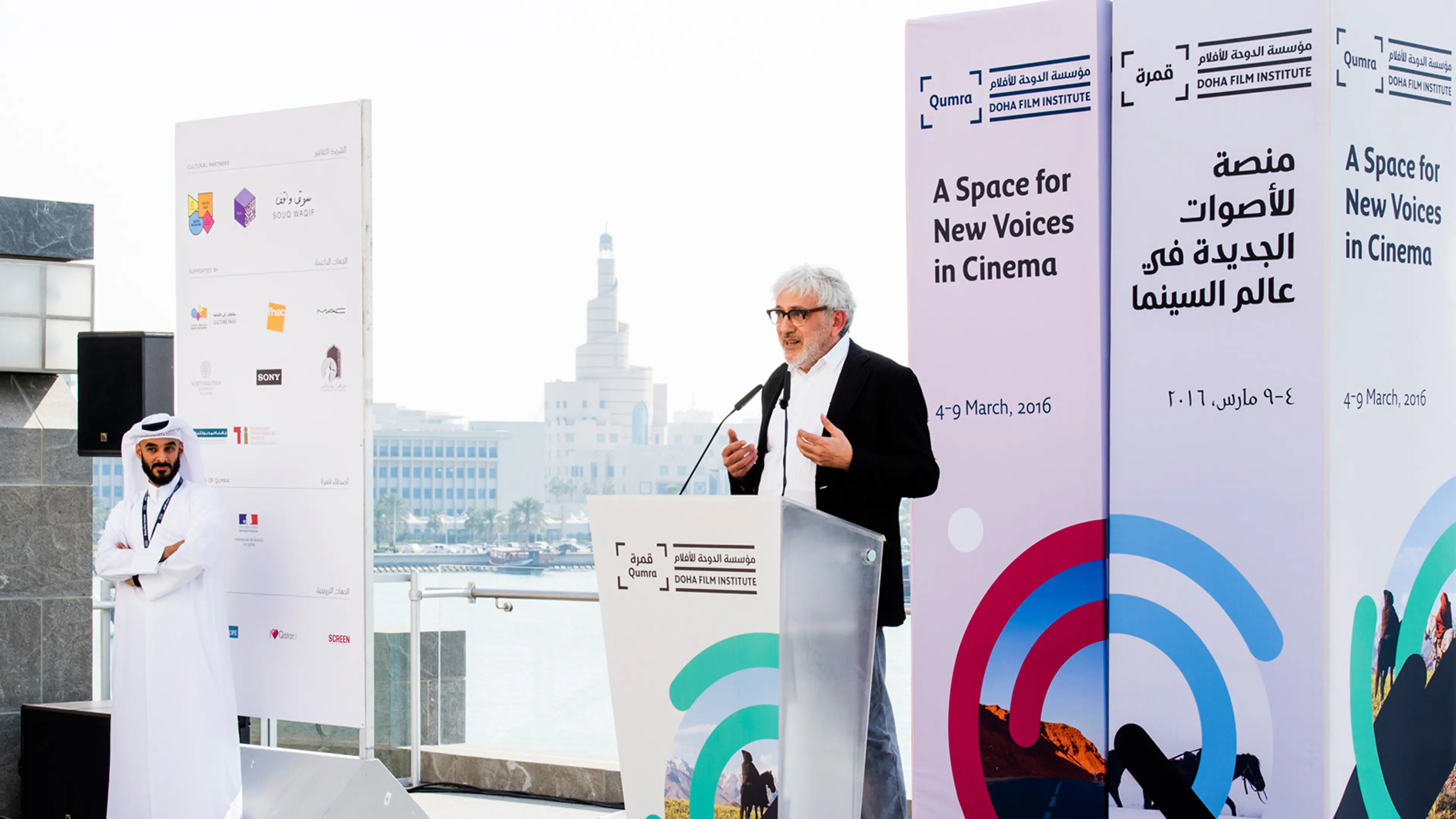 Man in black jacket speaking at a podium with Doha Film Institute banner reading 'A Space for New Voices in Cinema' and a man in traditional white attire standing with arms crossed.