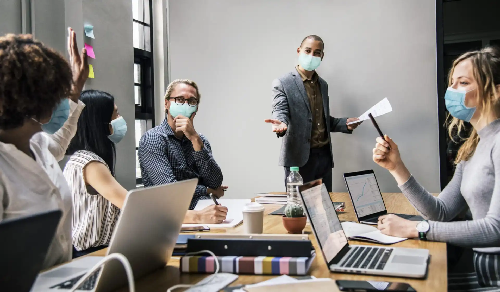 Reunião de equipe em um ambiente corporativo, com participantes usando máscaras e tecnologia como laptops e mesas de trabalho, destacando a colaboração e o trabalho em equipe.