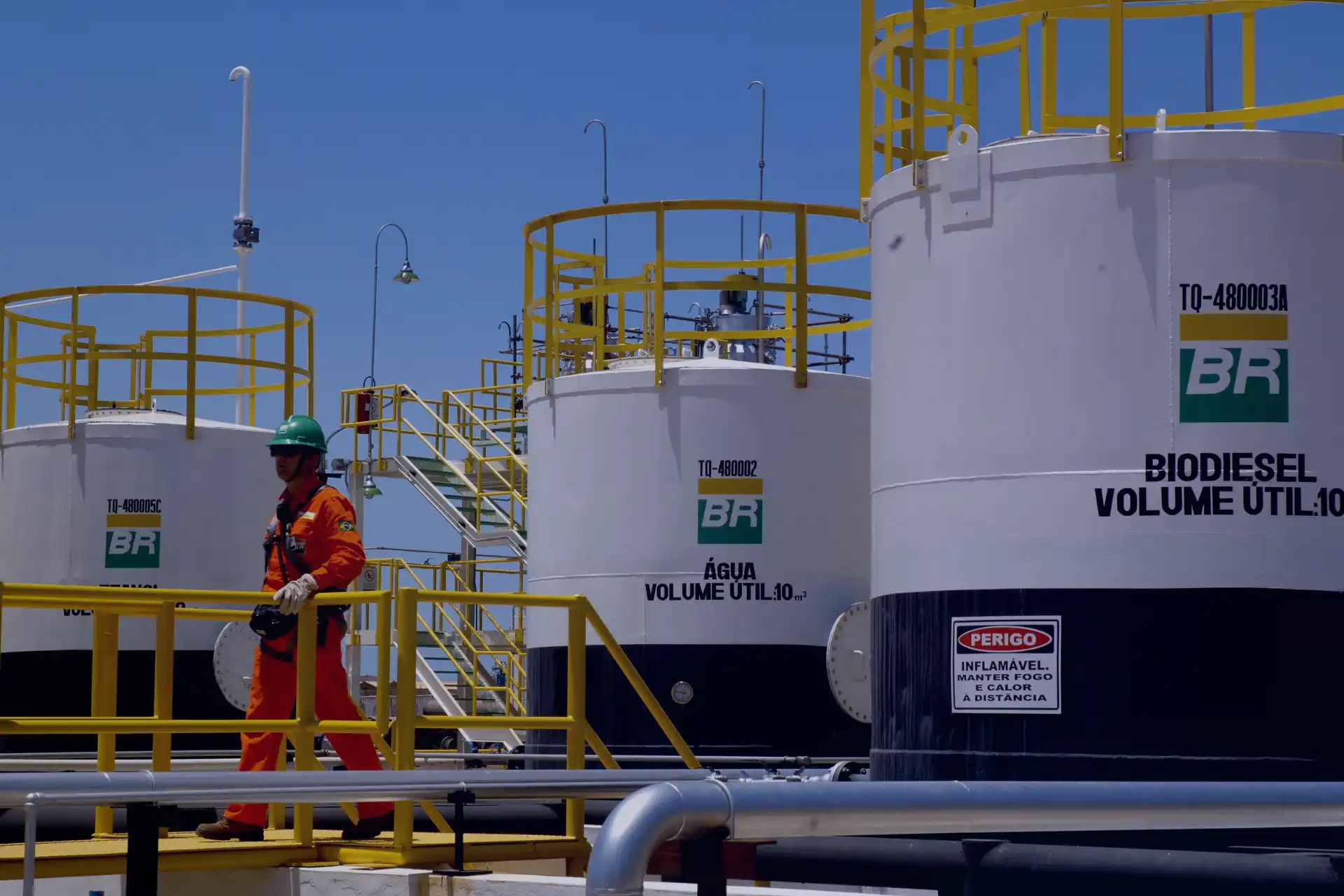 Tanques de biodiesel e água em instalação industrial com trabalhador usando equipamento de proteção. Imagem representa a indústria de energia renovável.
