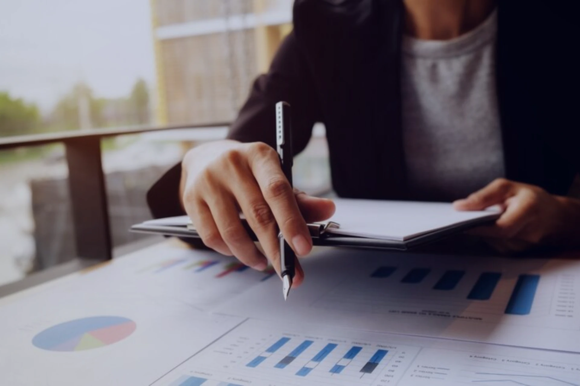 Pessoa analisando gráficos e relatórios financeiros em um ambiente de trabalho. A imagem mostra uma mão segurando um caderno e uma caneta em um escritório moderno.