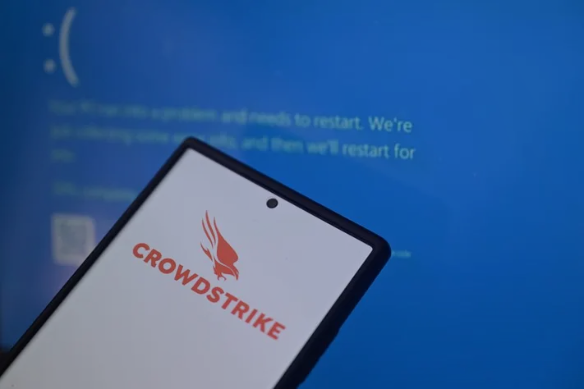 Imagem de um celular exibindo o logotipo da CrowdStrike, em frente a uma tela de computador com mensagens de erro e reinicialização.