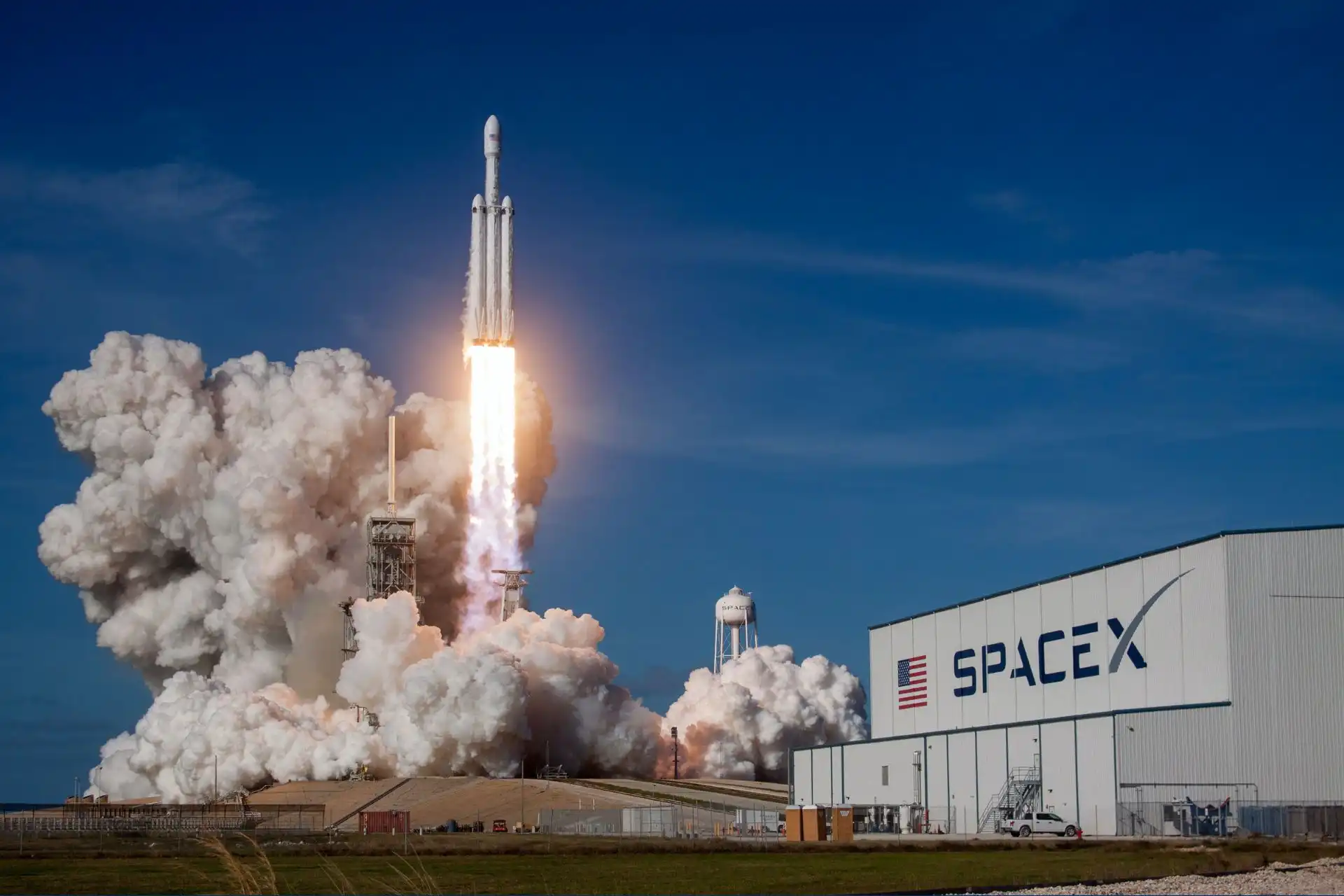 Foguete da SpaceX em lançamento, gerando uma nuvem de fumaça branca sob um céu azul, com a logo da SpaceX em destaque.