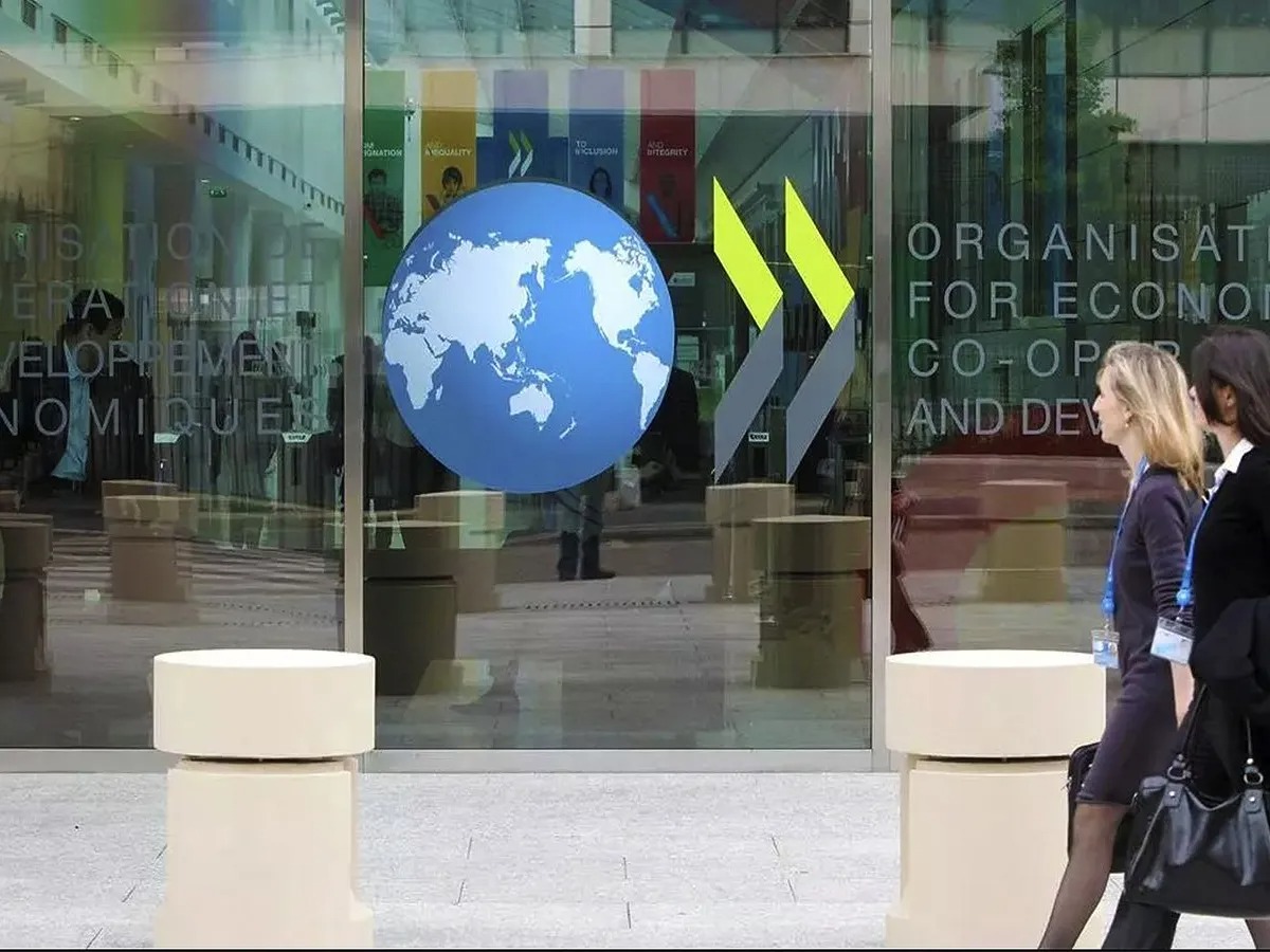 Imagem da fachada da Organização para a Cooperação e Desenvolvimento Econômico (OCDE) com um globo terrestre e pessoas caminhando.
