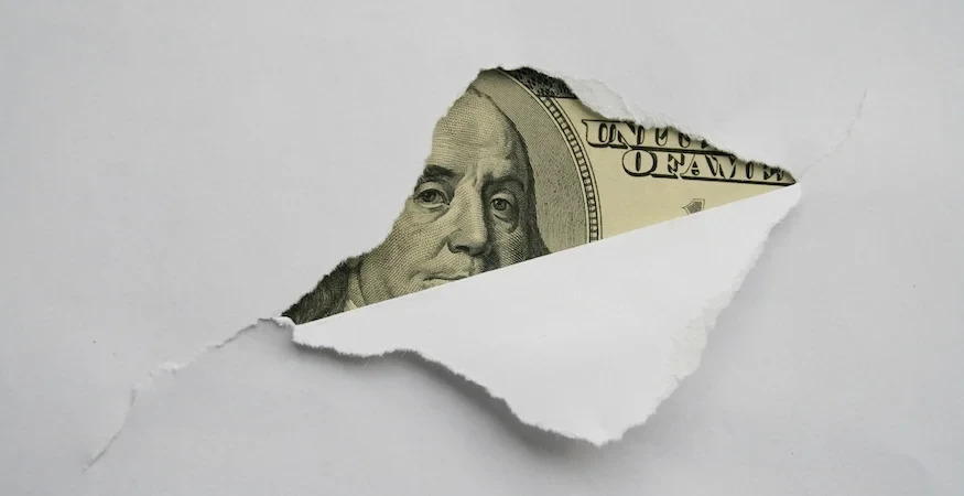 Imagem de nota de cem dólares com rosto de Benjamin Franklin visível através de um papel rasgado, simbolizando riqueza e oportunidades financeiras.