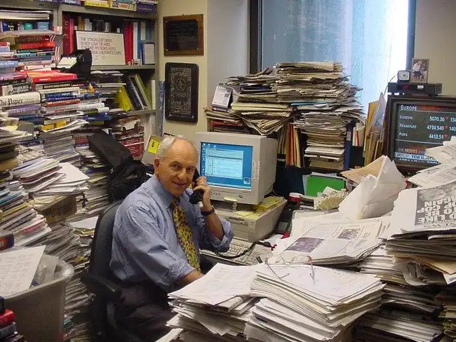 Homem em um escritório desordenado, cercado de pilhas de papéis e livros, falando ao telefone. Ambientes caóticos são comuns em escritórios de trabalho intenso.