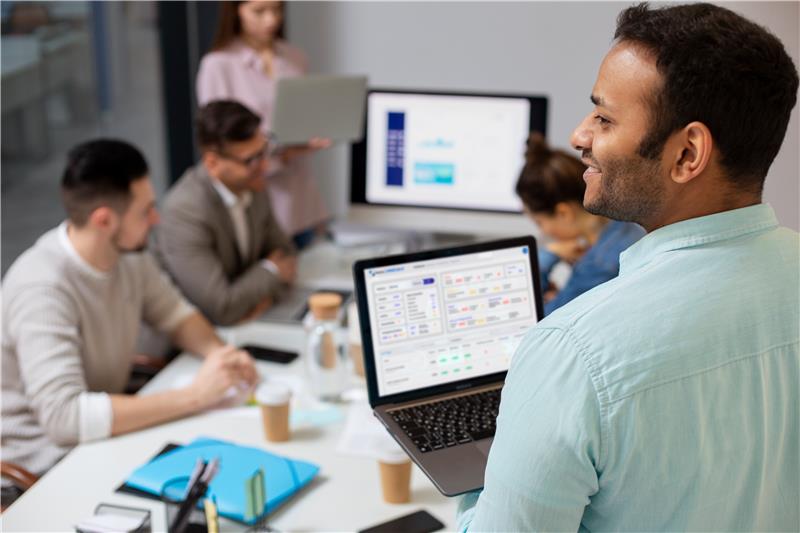 Homem sorridente em reunião de negócios com um laptop, mostrando apresentação de dados enquanto colegas discutem estratégias em um ambiente corporativo.