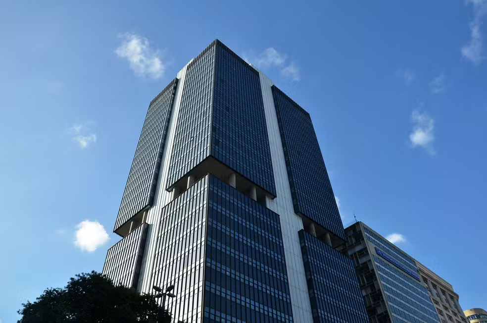Imagem de um prédio alto com fachada de vidro sob um céu azul, representando a arquitetura moderna. Este edifício é um exemplo do urbanismo contemporâneo nas grandes cidades.