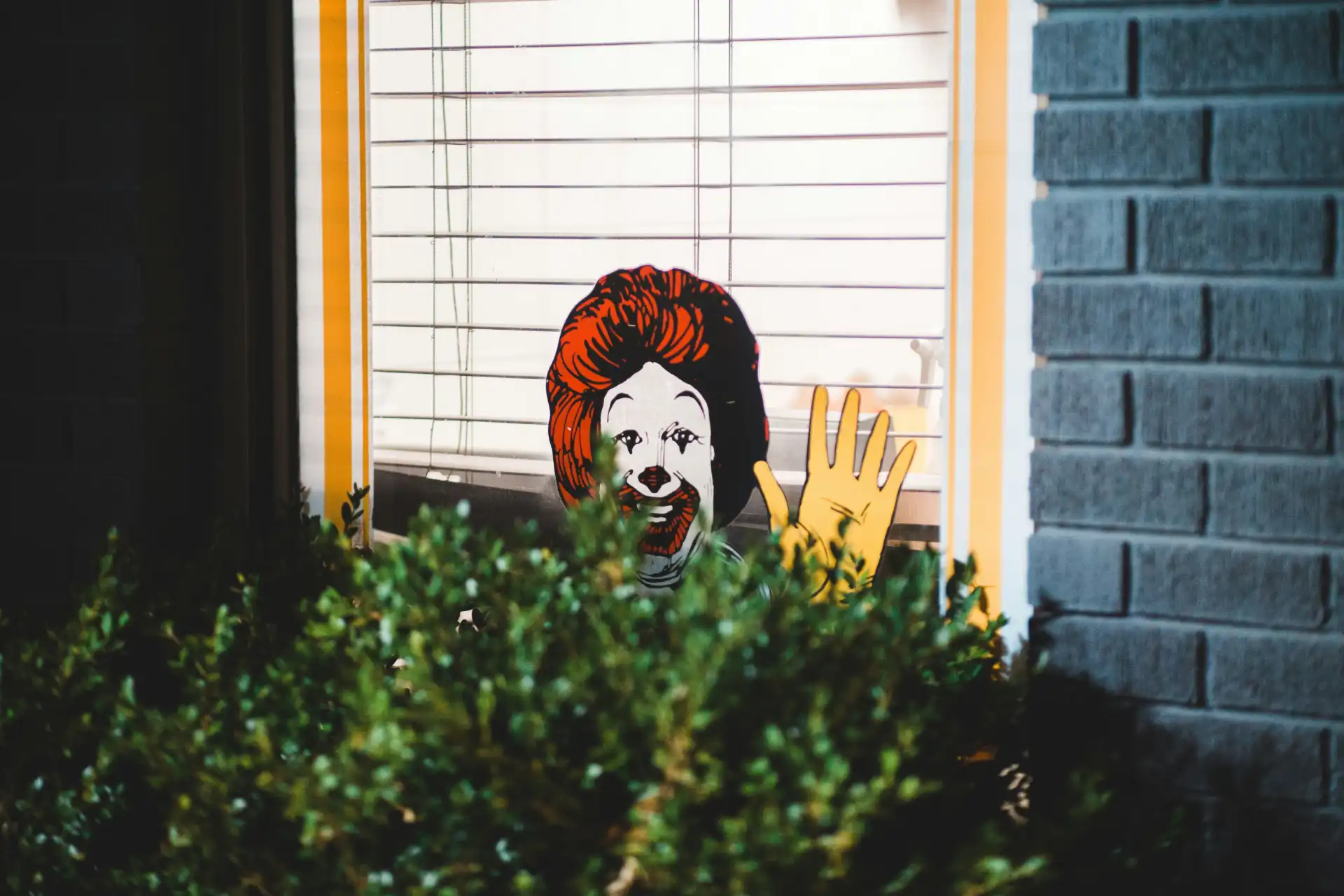 Imagem de uma placa decorativa com uma cara de palhaço assoviando, vendo pela janela, com plantas na frente e uma parede de tijolos ao lado.