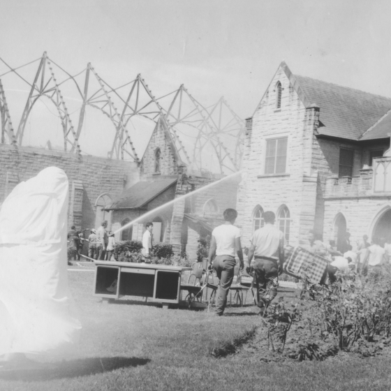 A black and white photo of a partially destroyed gothic style chapel. The roof is missing and only framing is left in poor condition. There are walls intact, although they appear to have soot and burn marks. A different portion of the building appears to be undamaged.
There is a group of firefighters using large streams of fire hose water to put out the remaining fires. There are also groups of people who appear to be assisting with the removal of furniture.