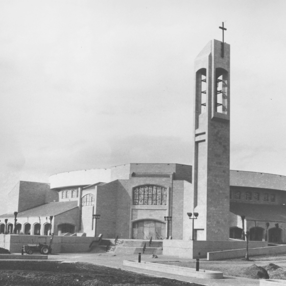 A black and white photo of the rebuilt Church Basilica. A large steeple, twice the height of the building, hangs over the structure complete with a cross on top. The church building has a large open stairway leading up to the entry doors. There is a large vehicle on site as part of the construction activity.