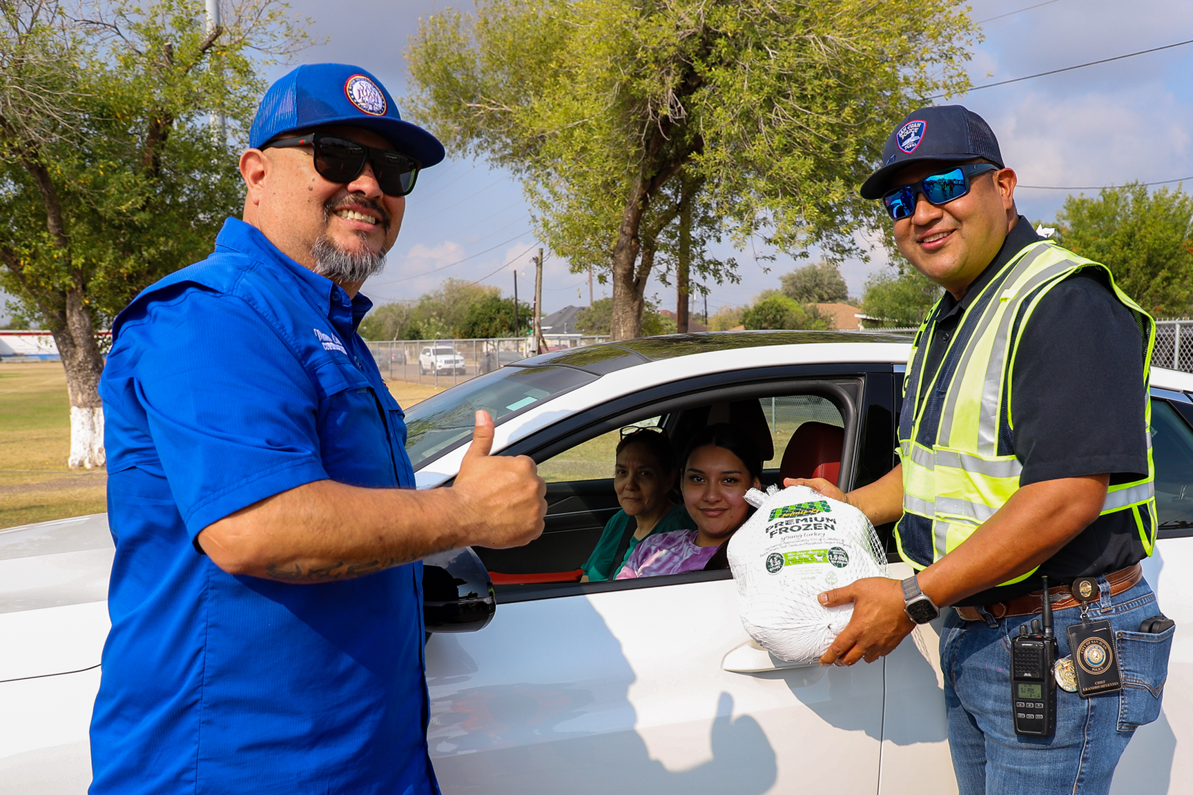 Two men wearing sunglasses, one in a blue shirt giving a thumbs up and the other in a reflective vest holding a frozen turkey, stand next to a white car with two women sitting inside.